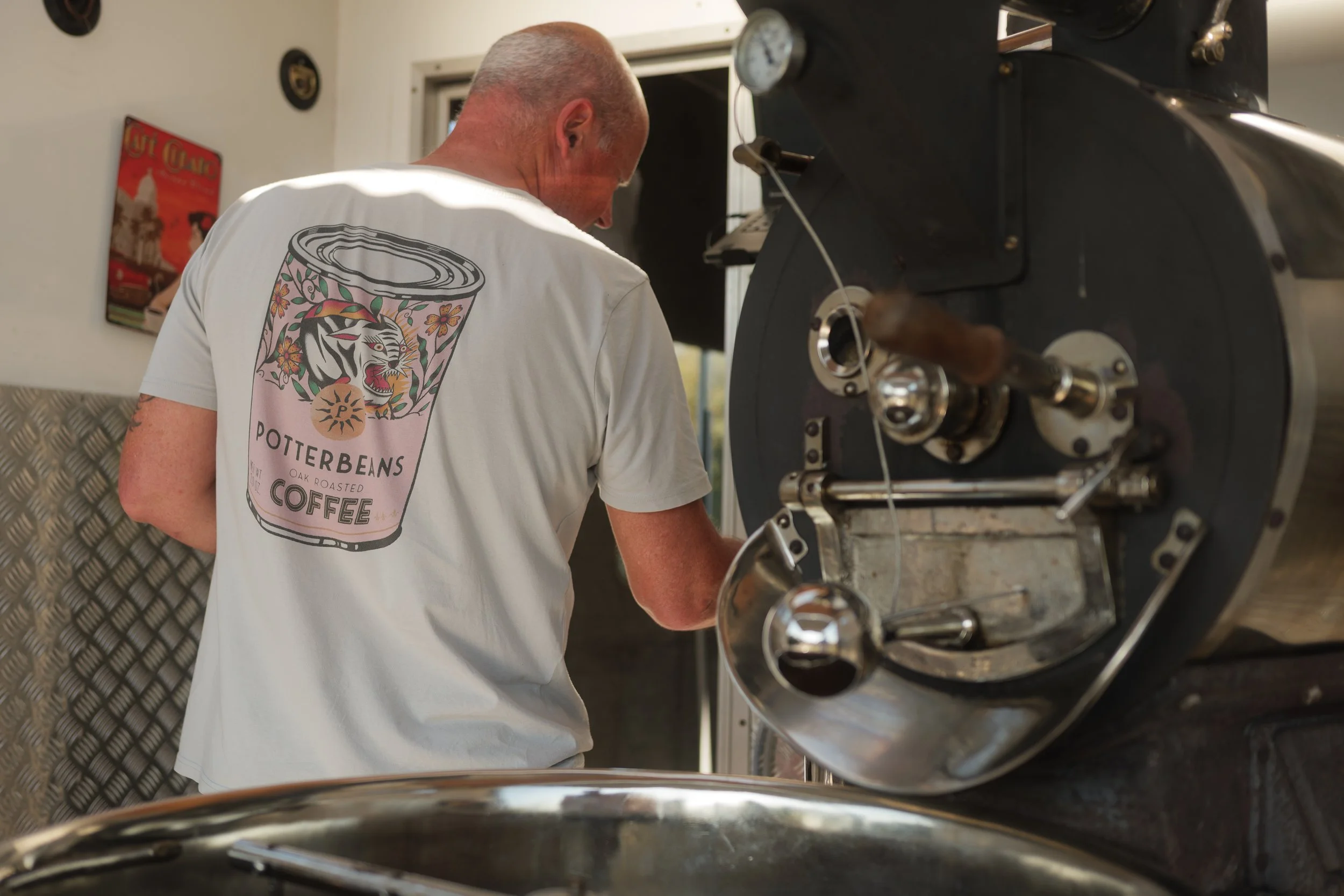 A man operating a machine, wearing a white t-shirt with a Pottery Barn coffee can design on the back, in a room with a white wall and a framed poster.