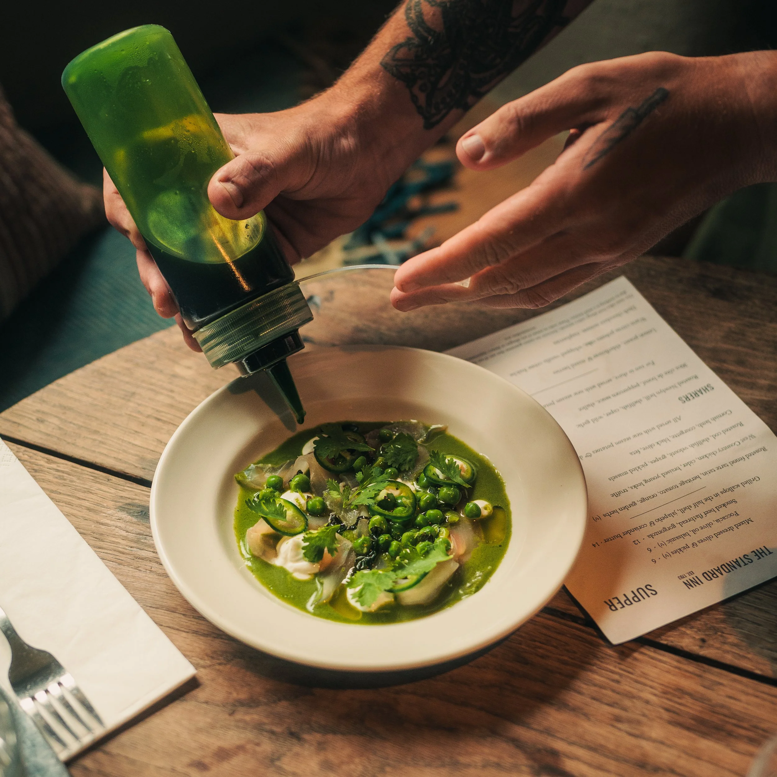 Person pouring a green sauce or dressing from a squeeze bottle onto a bowl of food, garnished with cilantro and peas, on a wooden table with a printed menu or paper nearby.