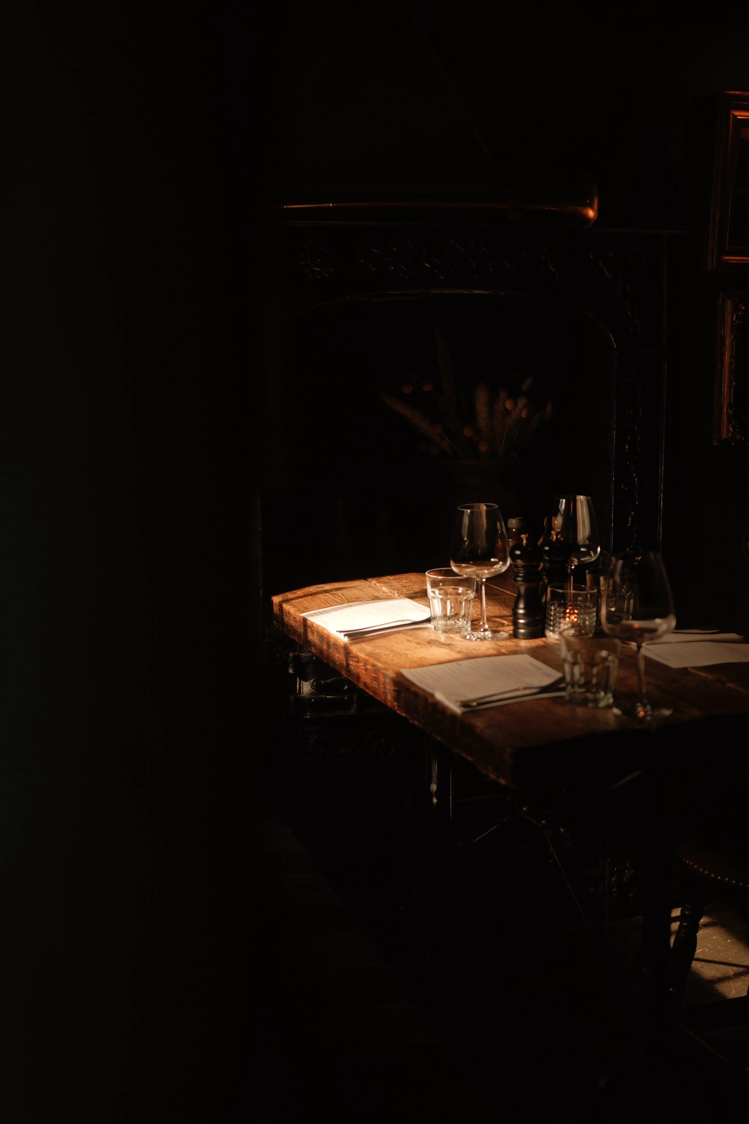A dimly lit dining table set with glasses, knives, and napkins in a dark room with a decorative mirror and frame on the wall.