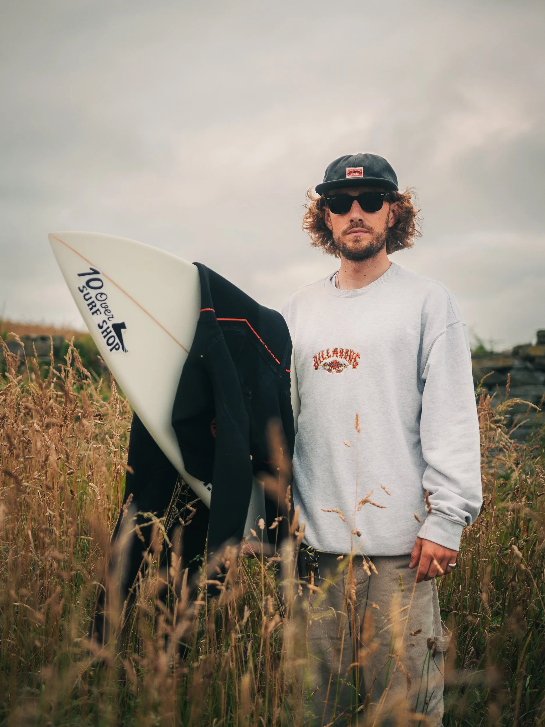A man standing in a field of tall grasses, holding a surfboard, wearing sunglasses, a black cap, a light gray sweatshirt, and beige pants.