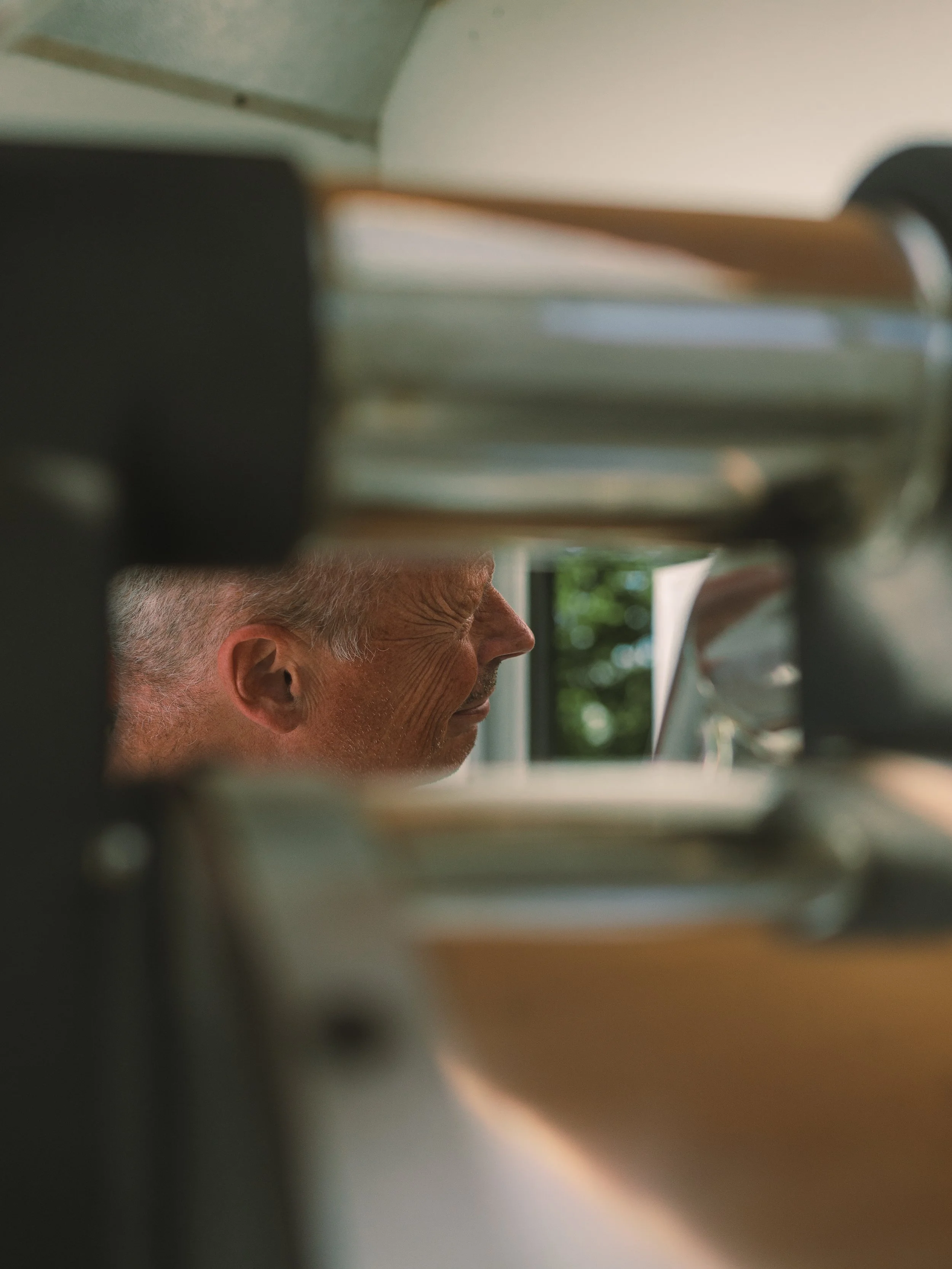 A close-up view of a person's face, likely a man, partially obscured by a blurry foreground element, with a background of a window showing green trees outside.