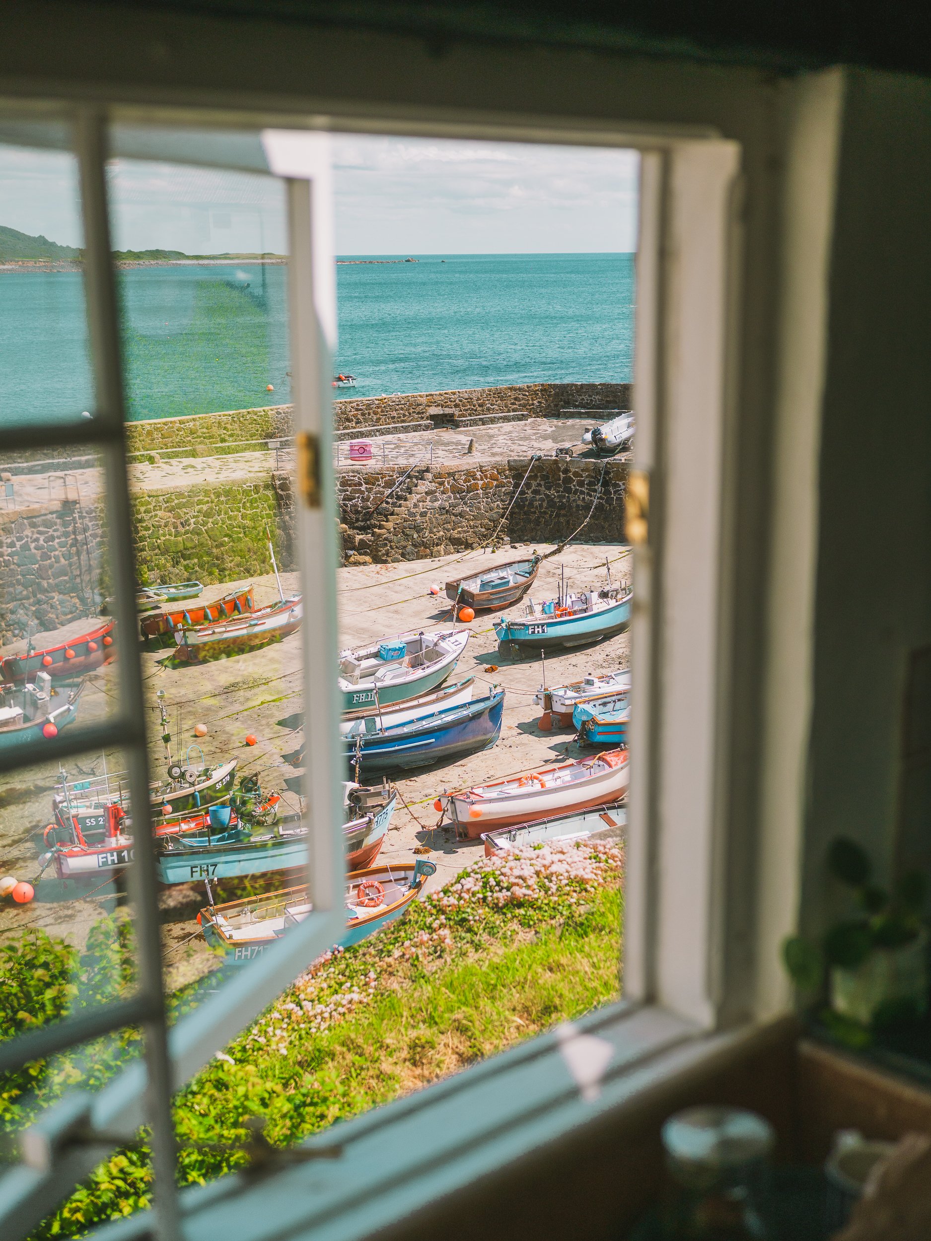 View of a small harbor with boats docked on a sandy shore, viewed through a window with a white frame. The sea is turquoise, and the sky is partly cloudy.