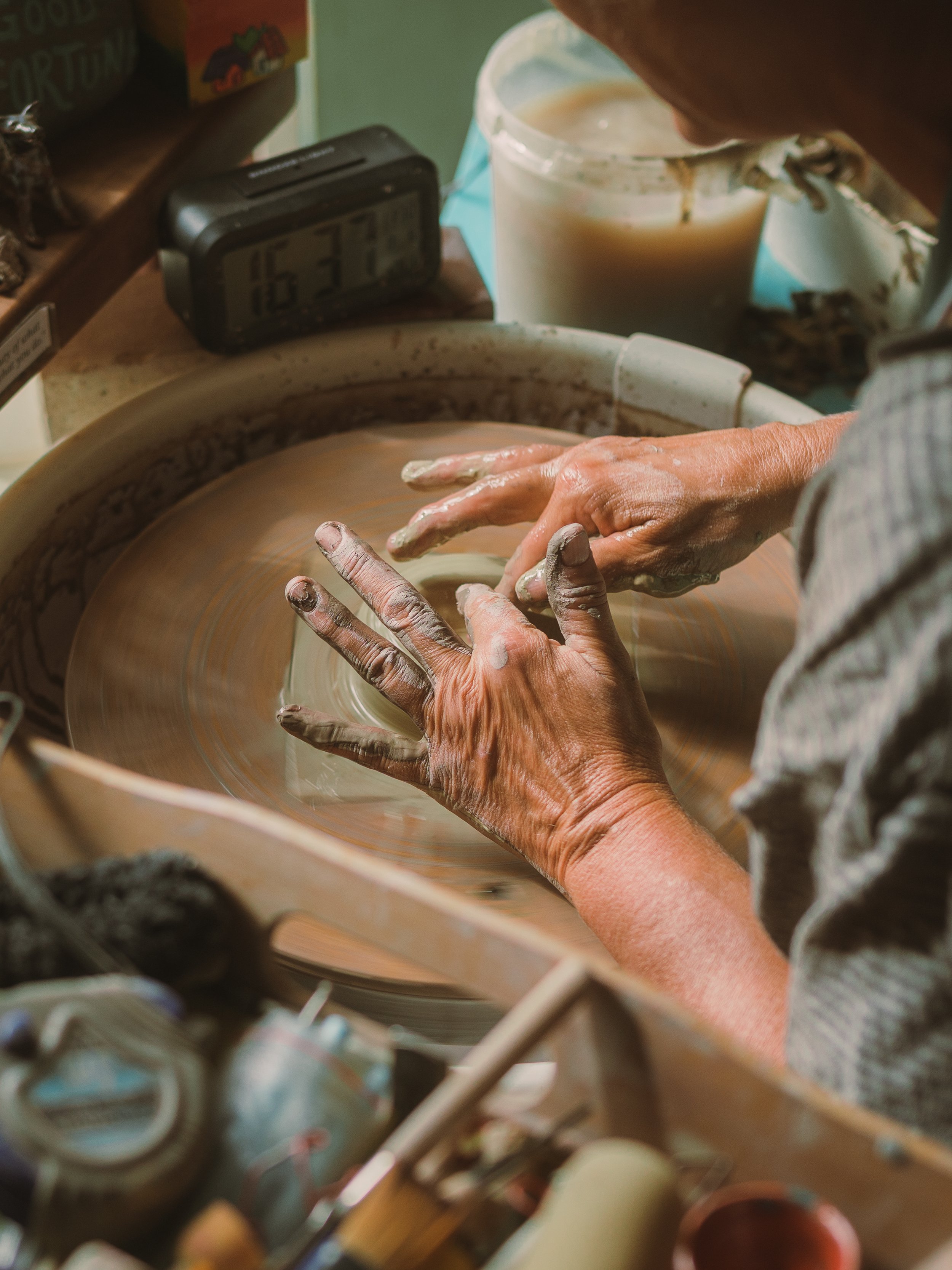 A person is shaping a ceramic piece on a pottery wheel with mud-covered hands, surrounded by pottery tools and a container with water or slip.