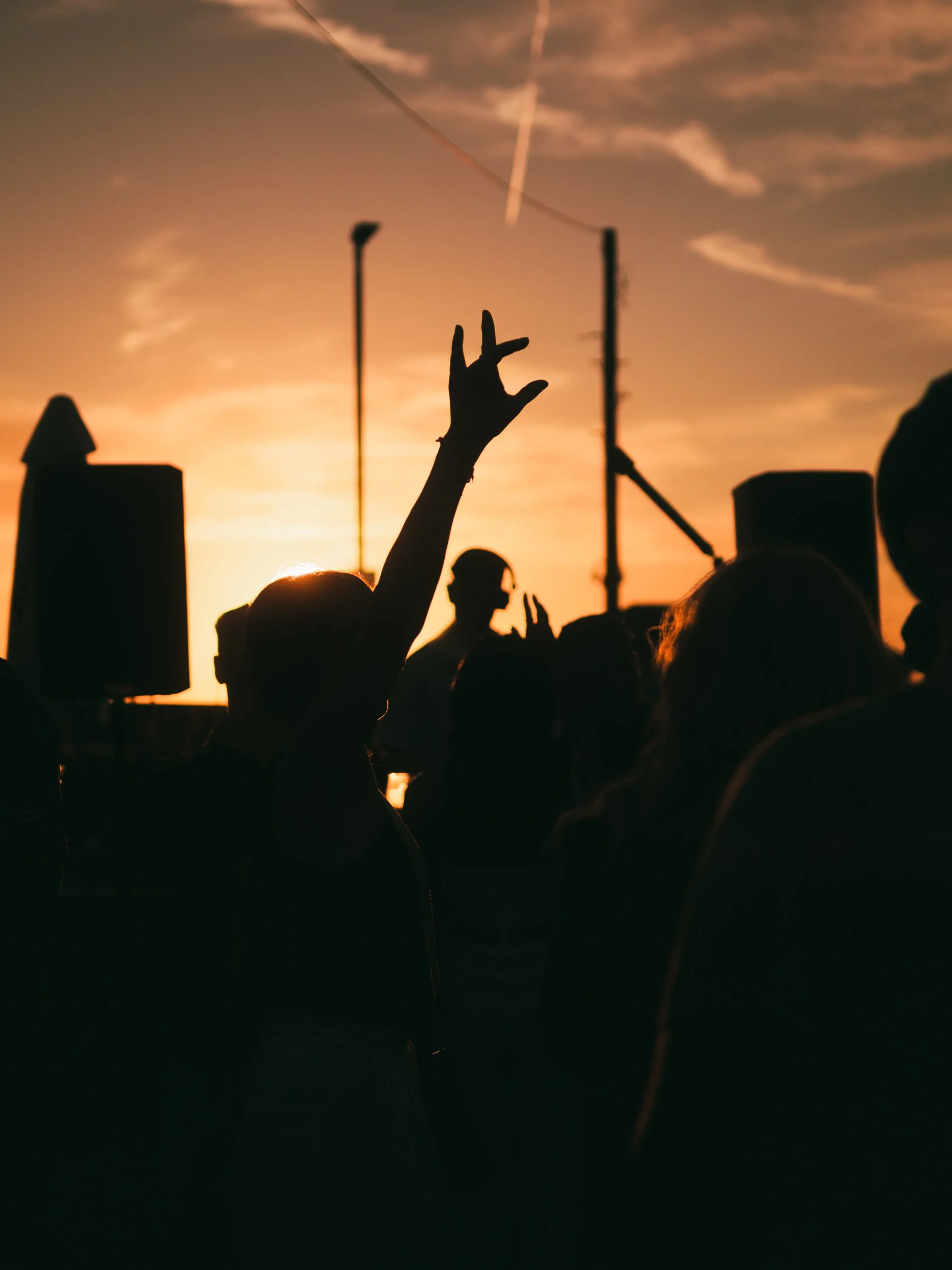 Silhouettes of several people at a concert or event during sunset or twilight, one person raising their hand in a rocker gesture, with stage equipment and a colorful sky with clouds and contrails in the background.