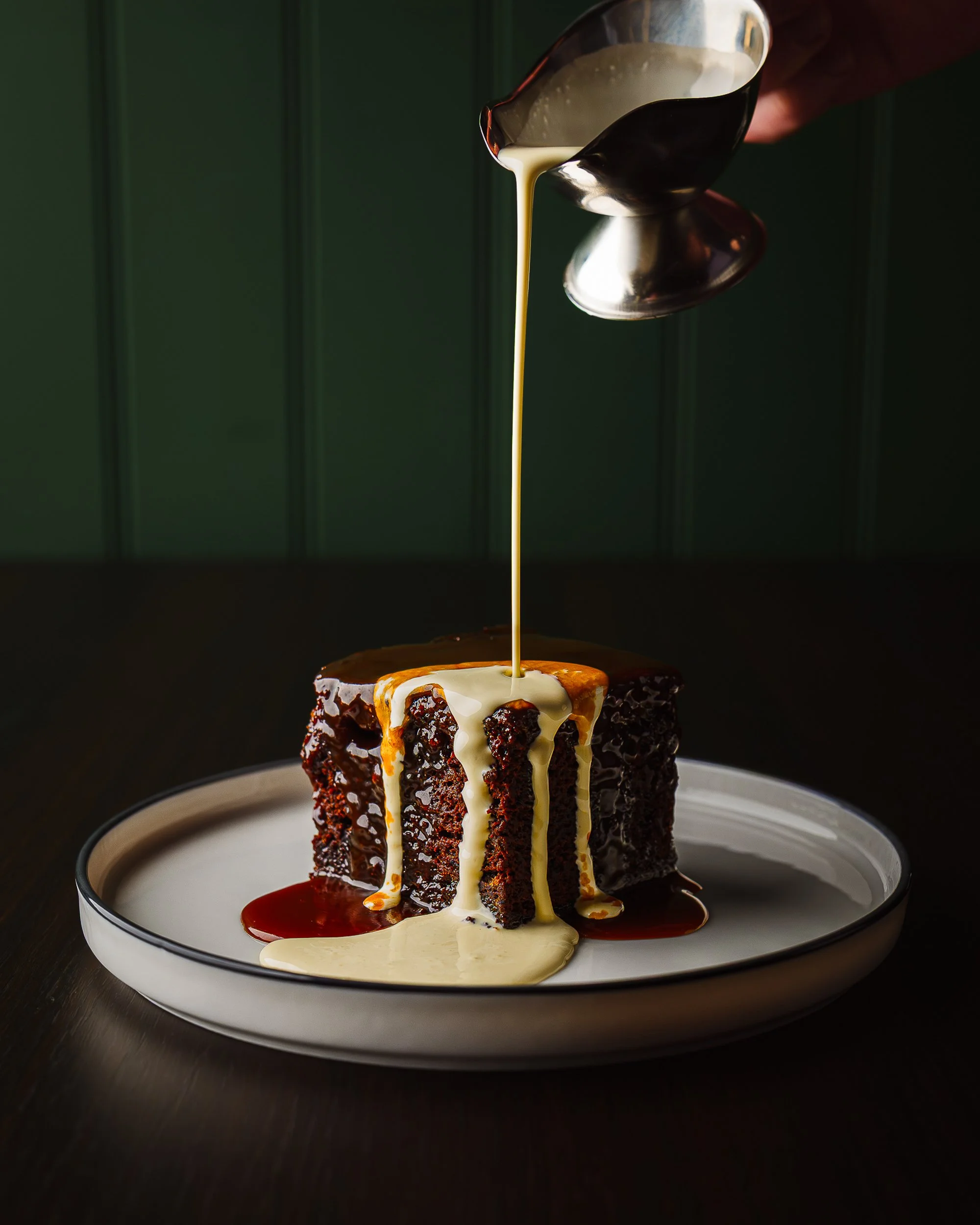 A piece of chocolate cake with caramel and white sauce on a white plate, being drizzled with cream or sauce from a metallic pouring vessel, against a dark background.