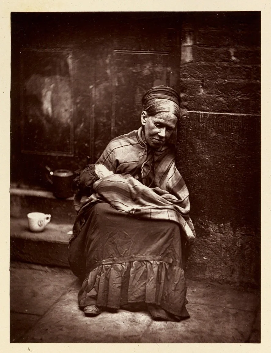 An elderly woman sitting on a step next to a brick wall, holding a sleeping baby wrapped in a blanket. There are two cups placed on the ground nearby.