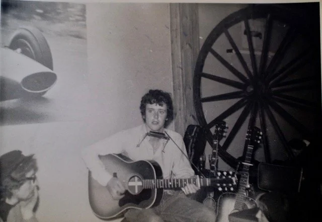 A young person playing an acoustic guitar with a harmonica holder, in a room with a large wooden wheel and several other guitars in the background.