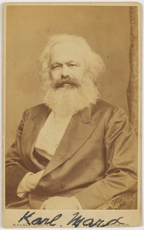 A sepia-toned photograph of an elderly man with a full beard and mustache, wearing a suit jacket and shirt, sitting with one hand resting on his lap and the other arm resting on the armrest of a chair. There is a handwritten signature reading "Karl Marx" at the bottom of the photograph.