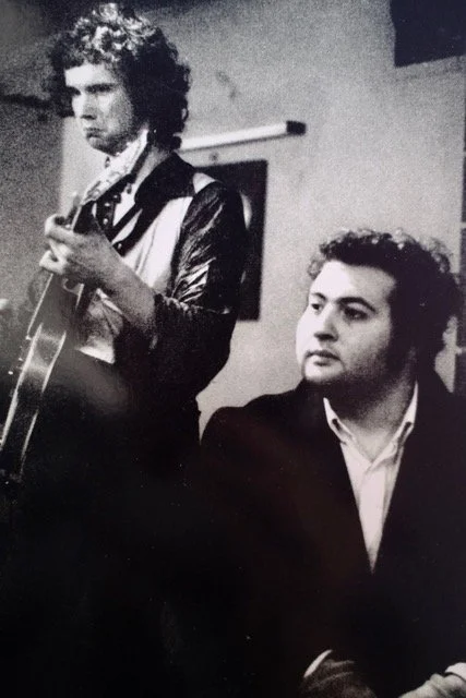 Black and white photo of two men, one playing a guitar and the other sitting nearby, indoors.