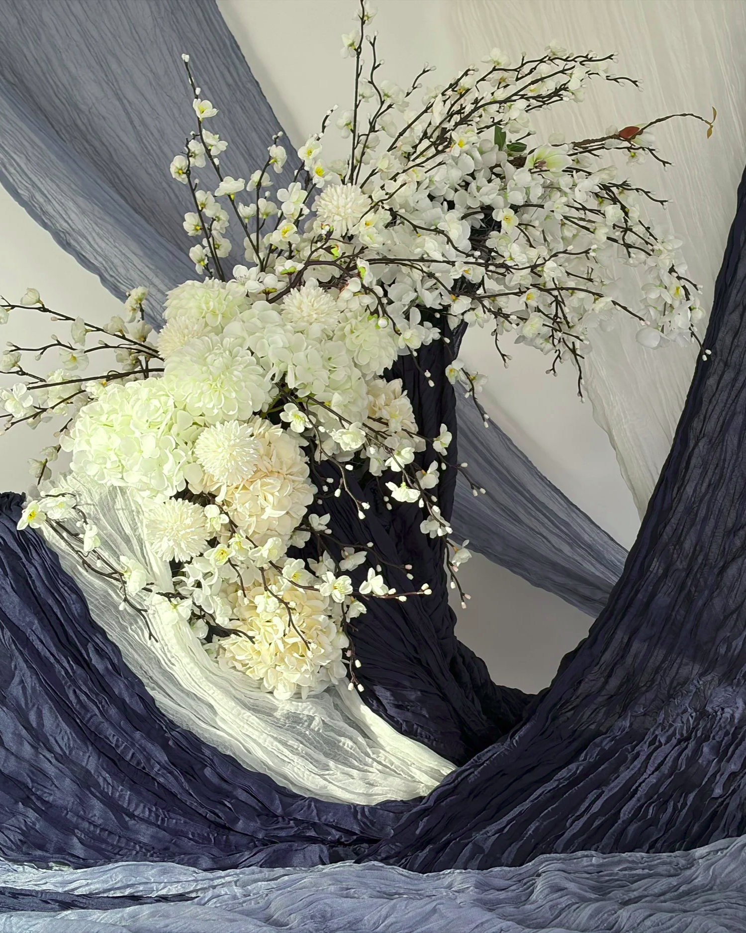 A floral arrangement with white hydrangeas, daisies, and branches with small buds is displayed inside a black and gray textured circular frame.
