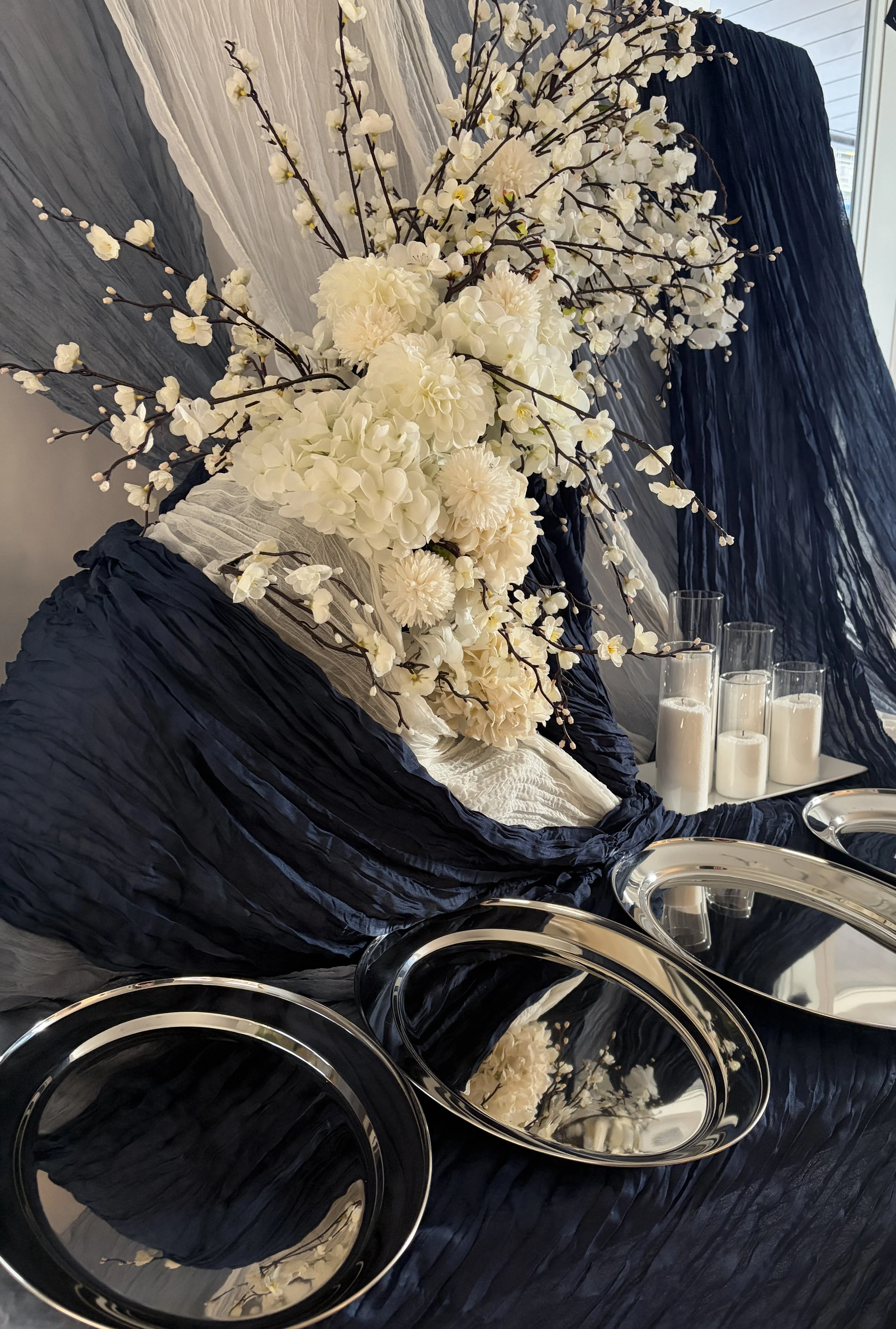 A large flower arrangement with white flowers and dark branches on a dark fabric backdrop with draped white fabric. Several tall glass candle holders with white candles are placed in front of the arrangement.