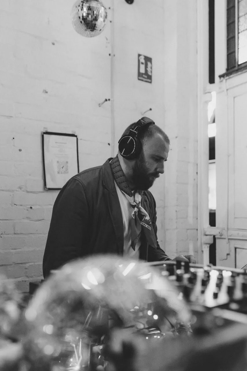 A man wearing headphones is working with electronic equipment in a room with white brick walls and large windows. A disco ball hangs from the ceiling.