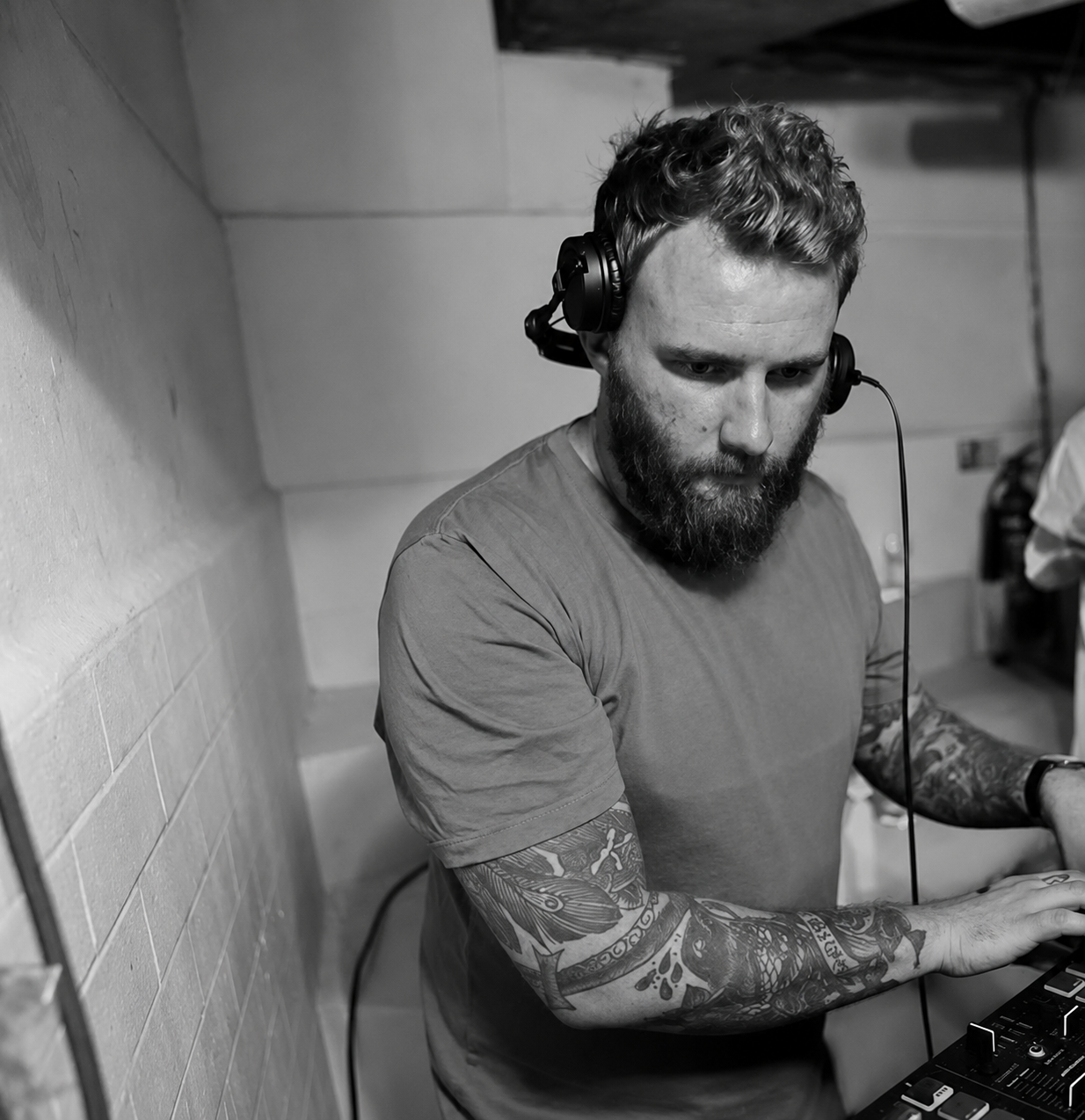 A man with a beard and tattoos wearing headphones is focused on a music mixer or DJ equipment in a room with brick and wooden walls.