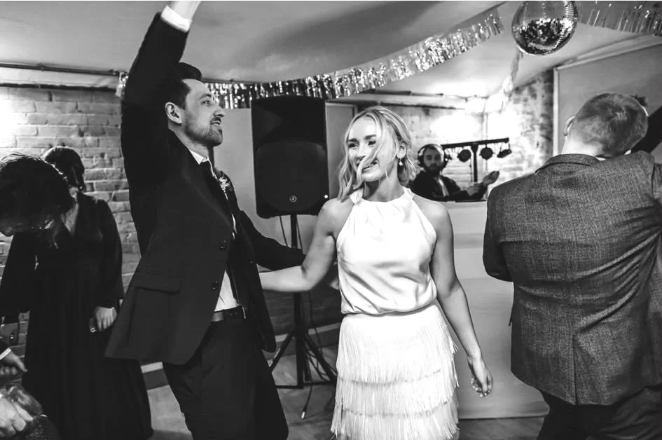Couple dancing at a celebration or wedding, with festive decorations and a DJ in the background.