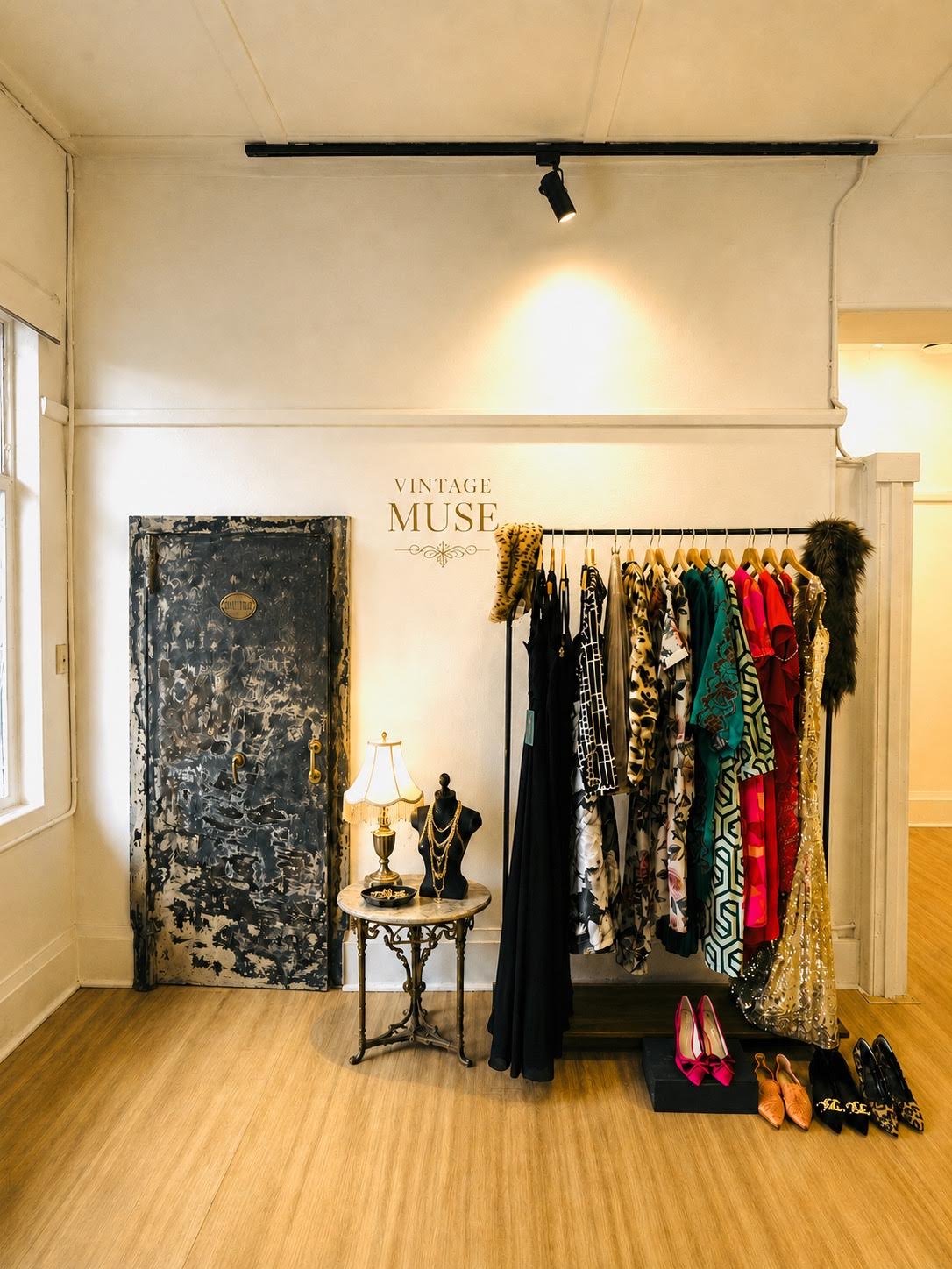 Vintage boutique display featuring a clothing rack with dresses and garments, a small ornate table with jewelry and a lamp, a decorative door, and the sign 'VINTAGE MUSE' on the wall.