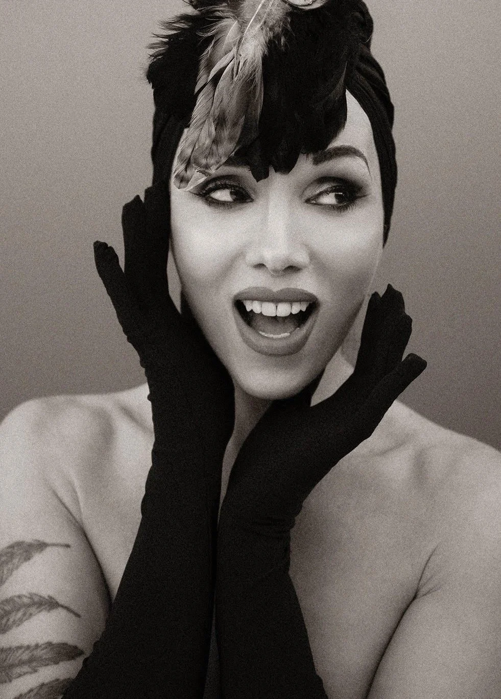A woman with dark hair wearing a feathered headpiece and long black gloves, posing with her hands framing her face, showing an expressive facial expression.