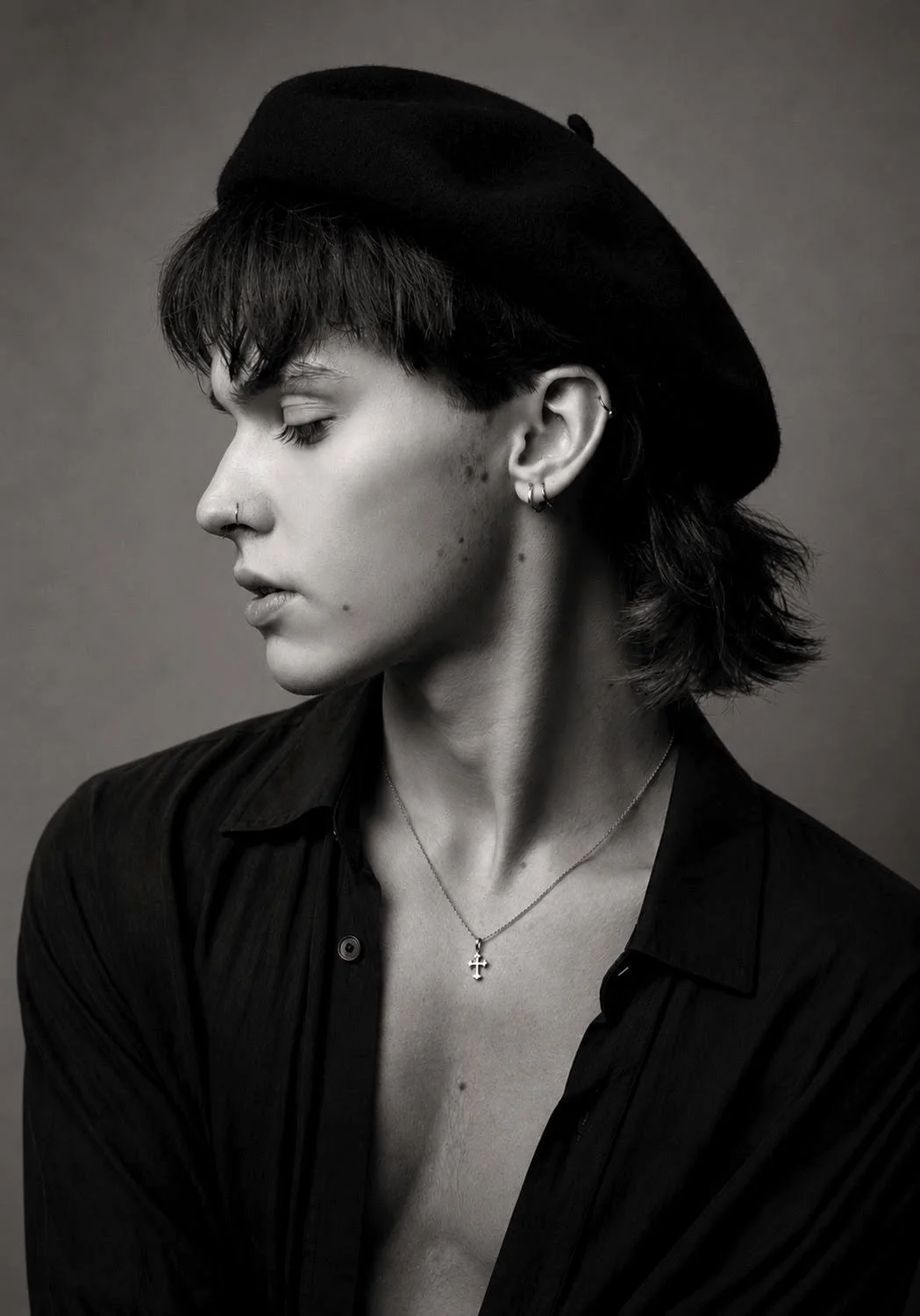 Black and white portrait of a person with short dark hair, wearing a black hat, multiple earrings, and a necklace with a cross pendant. They are dressed in a partially unbuttoned black shirt, looking to the side with closed eyes.
