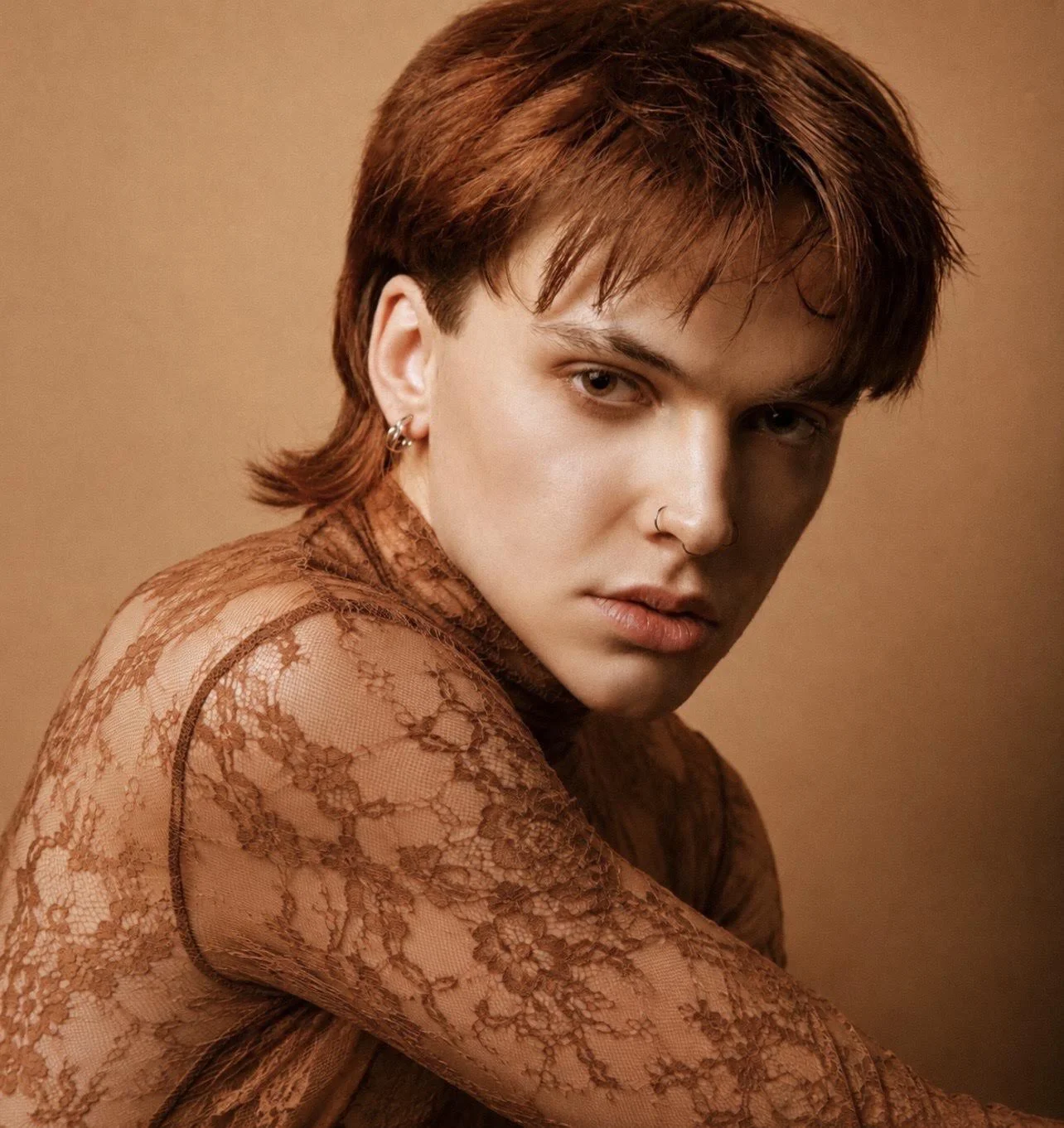 Person with short, reddish-brown hair, wearing earrings, a nose ring, and a brown lace top, posing against a plain background.
