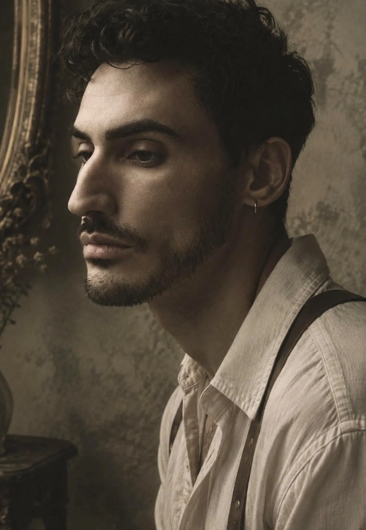 A close-up of a young man with dark curly hair, light beard, and earring, wearing a beige shirt with suspenders, looking to the side against a textured beige wall with an ornate picture frame.