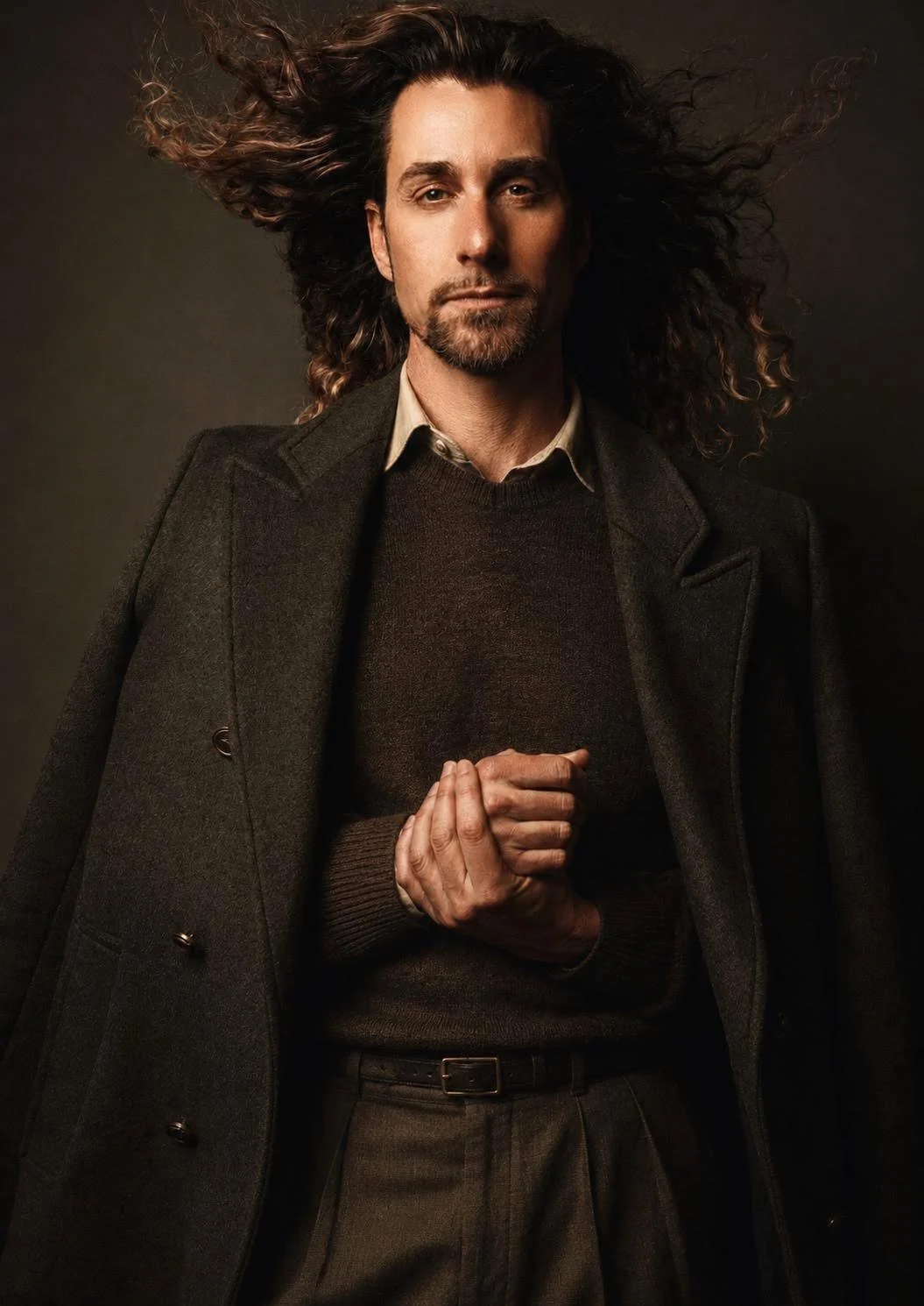 A man with long, curly hair and a beard wearing a dark blazer over a dark sweater and shirt, posing against a dark background.