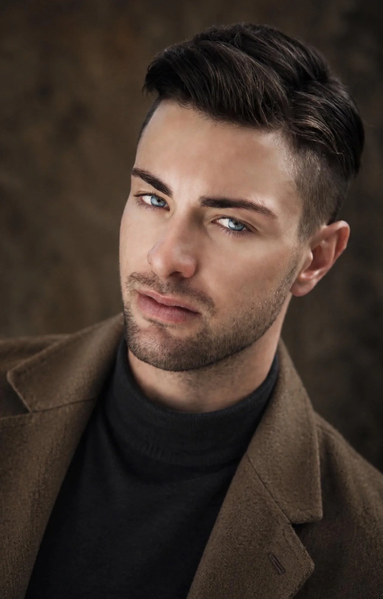 Close-up of a young man with styled dark brown hair, blue eyes, and light stubble, wearing a brown coat and black turtleneck against a blurred brown background.