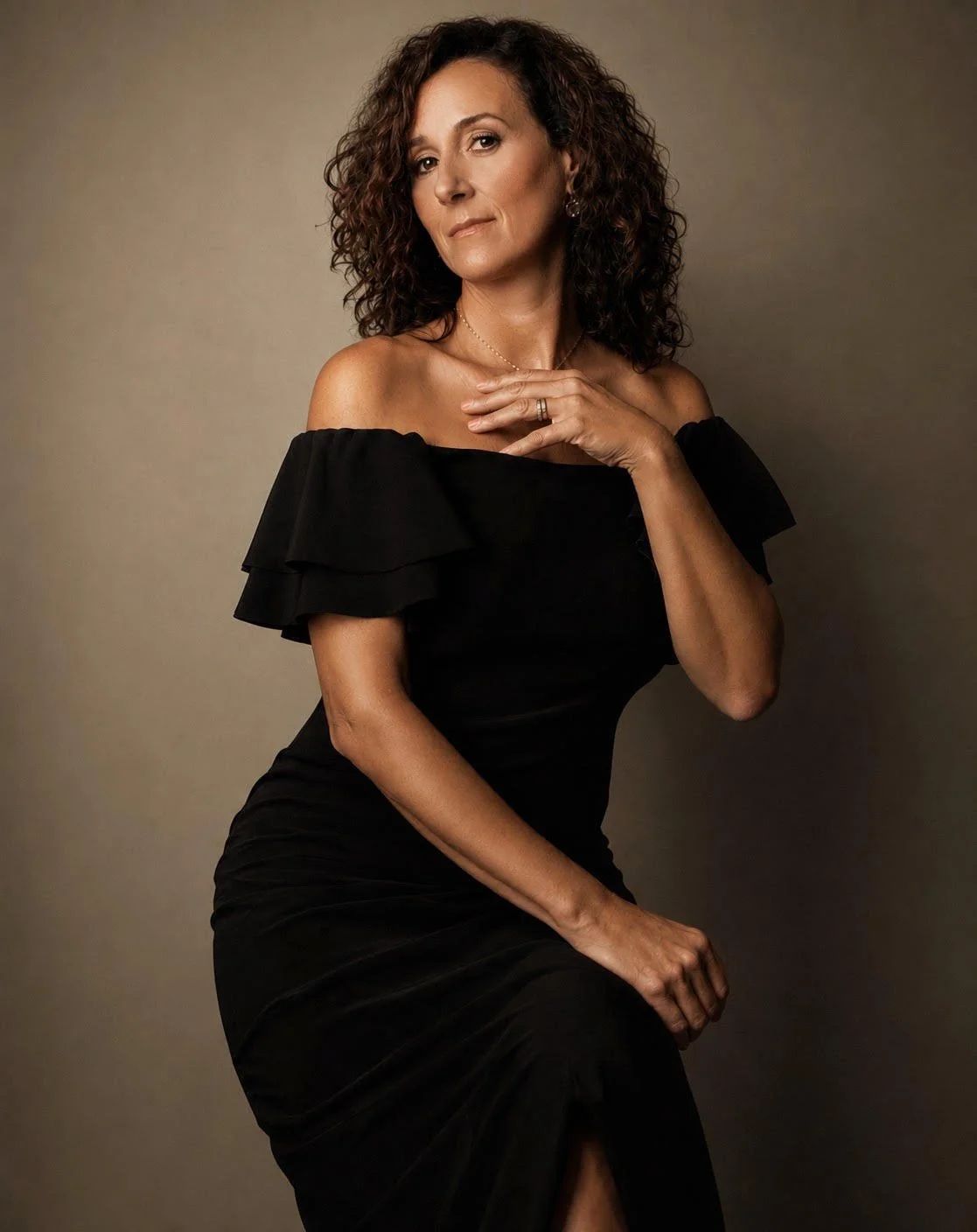 A woman with shoulder-length curly hair in a black off-the-shoulder dress, sitting against a plain background, looking confidently at the camera.