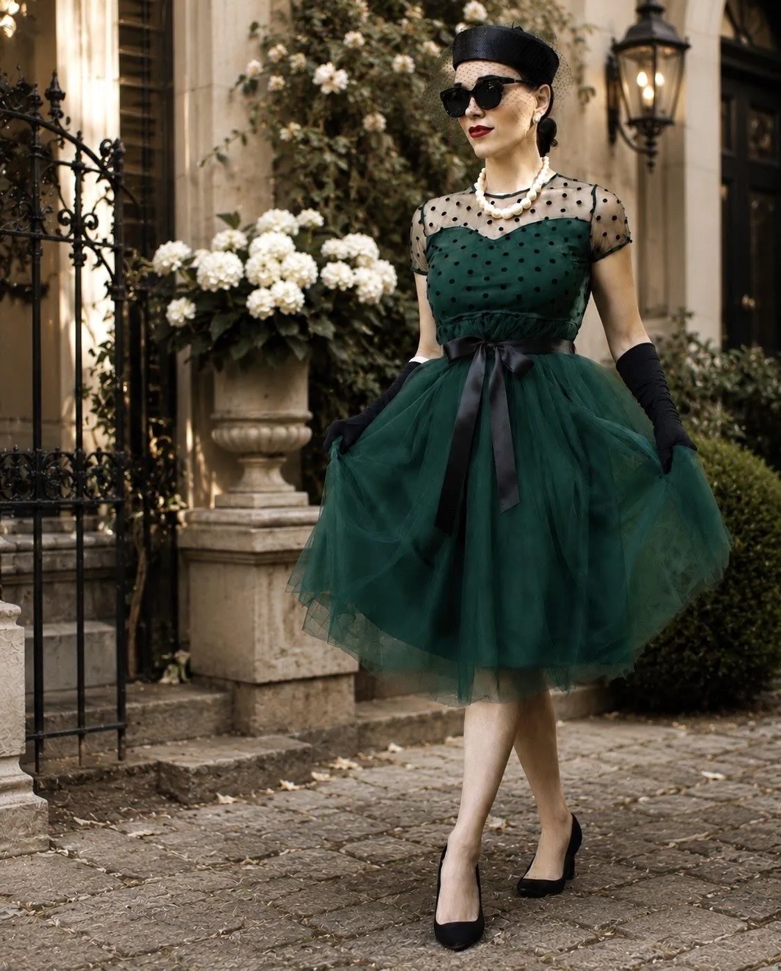 Woman in vintage style green dress with black polka dots, black hat, sunglasses, pearl necklace, black gloves, and high heels standing outdoors in front of a stone building with white flowers.