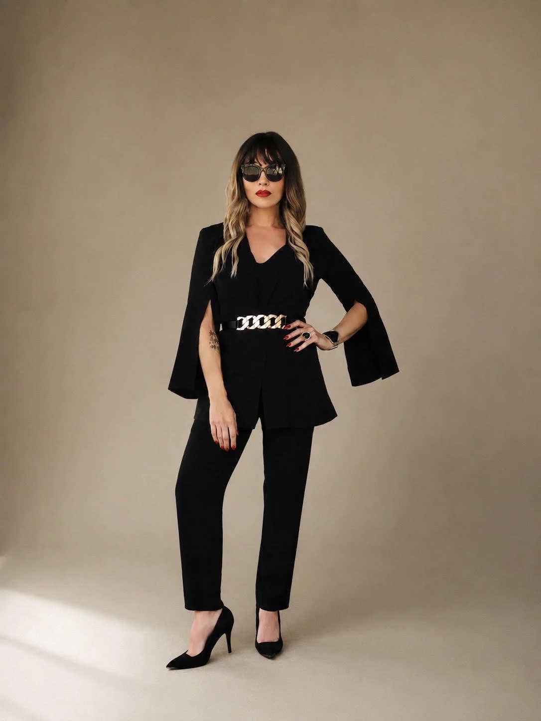 Woman in black suit with cape sleeves, black pants, black heels, sunglasses, red lipstick, and chain belt, standing against a plain beige background.