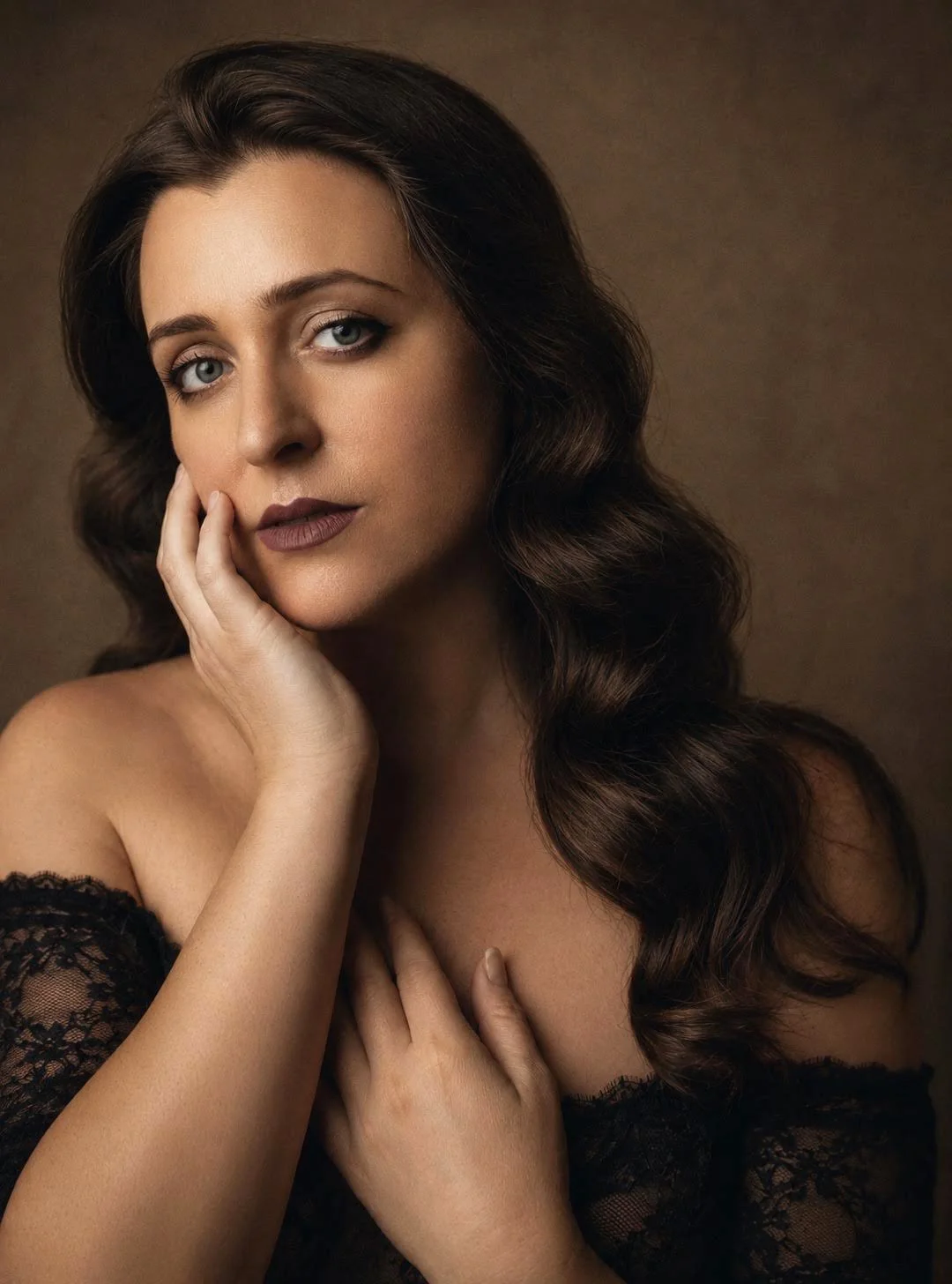 A portrait of a woman with long wavy dark hair, blue eyes, and fair skin, wearing a black lace off-shoulder top, posing with one hand on her face and the other on her chest against a brown background.