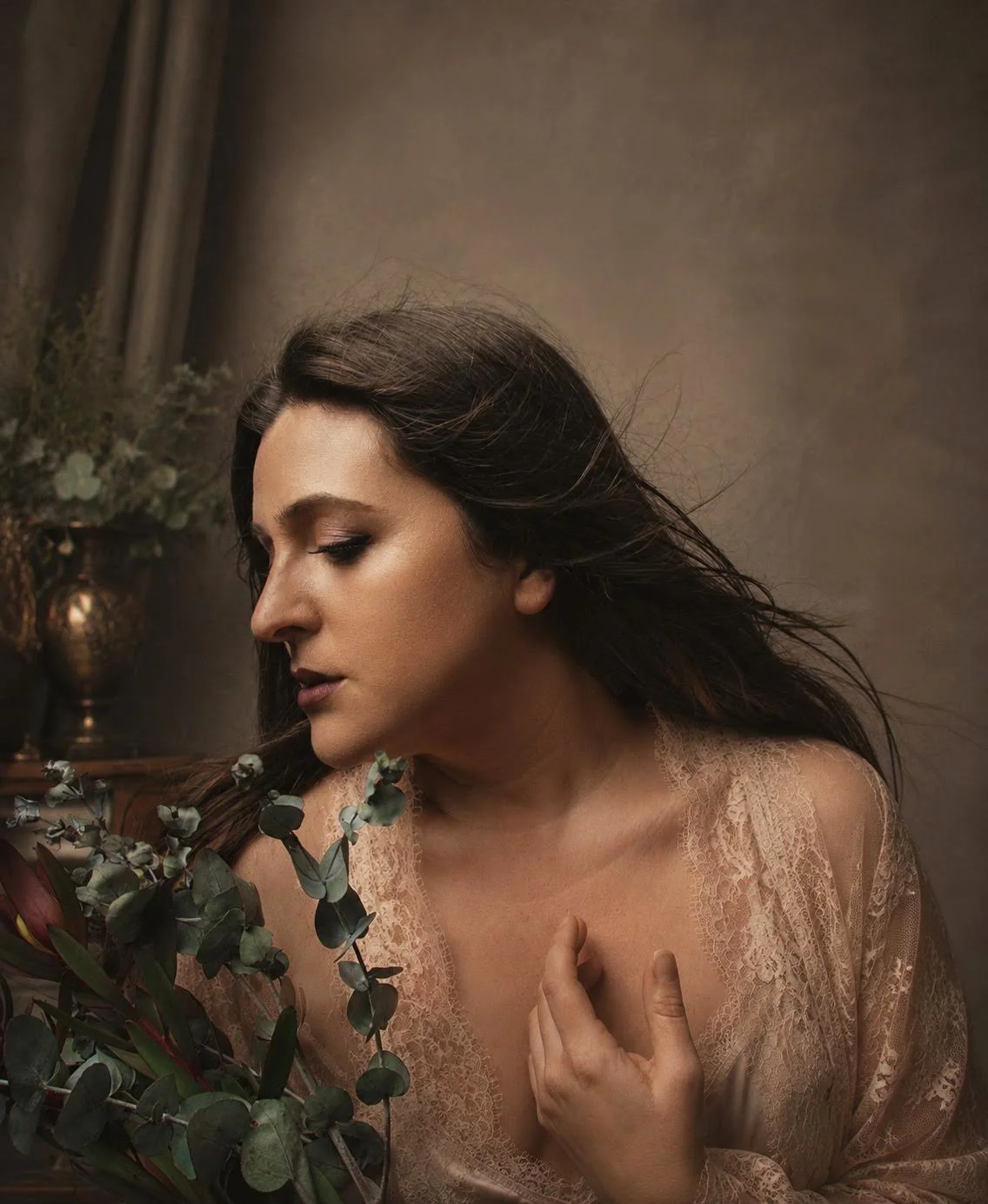 A woman with long dark hair and a lace top is close to a bouquet of eucalyptus and other flowers, with a soft, neutral background.
