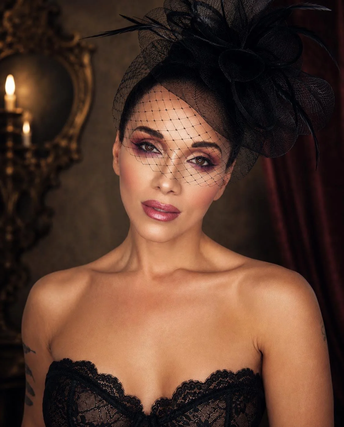 A woman with makeup and a black lace strapless top, wearing a black fascinator with a veil and feathers, in a dimly lit room with ornate decor.