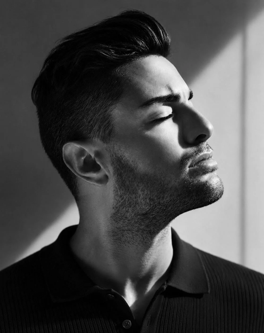 Black-and-white photo of a young man with closed eyes, styled hair, and facial hair, wearing a collared shirt, with sunlight casting a shadow on his face.
