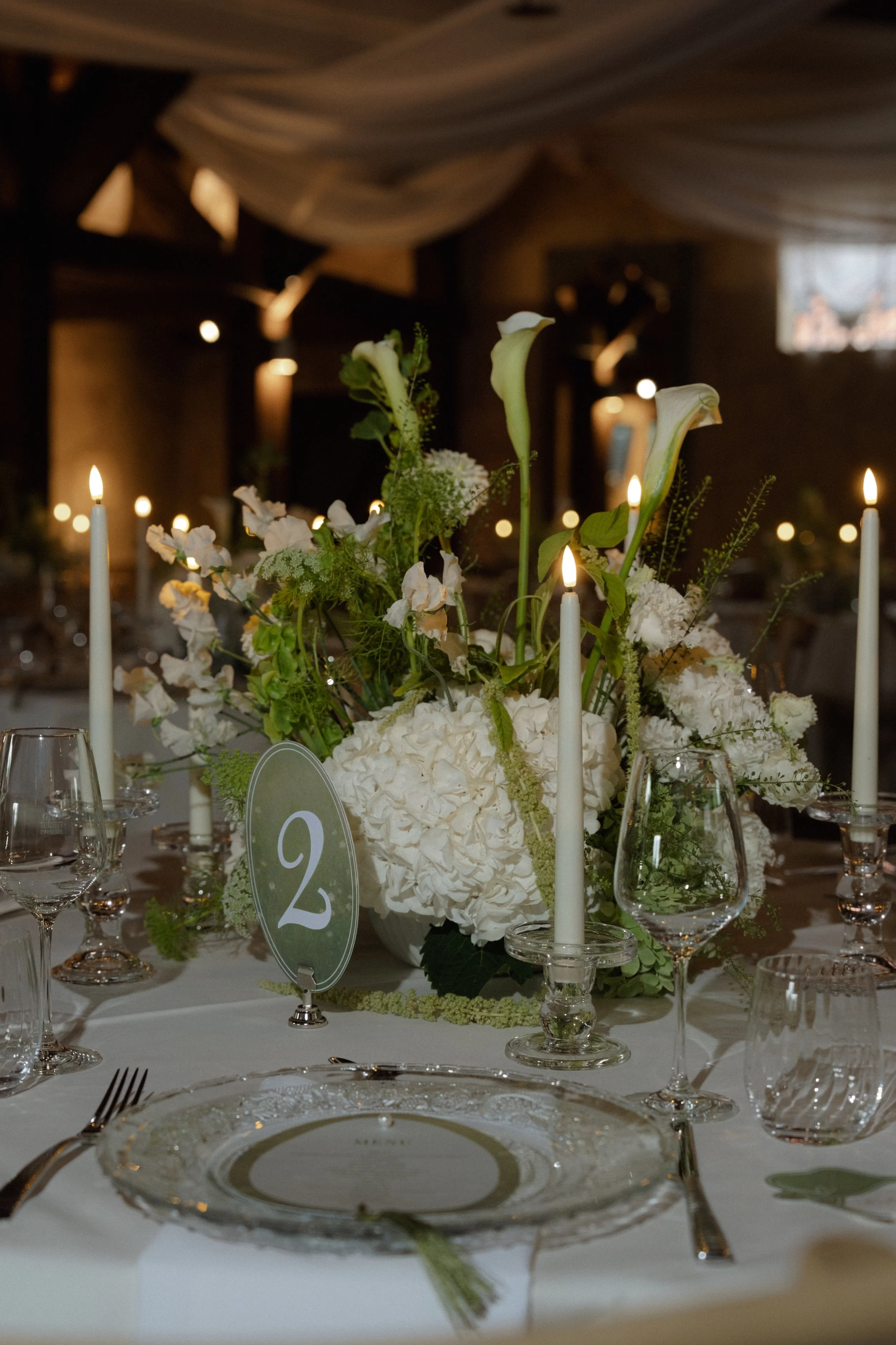 Man sieht einen eingedeckten Hochzeitstisch. Gläserne Platzteller, gläserne Kerzenhalter und ein Blumengesteck in der Mitte.
