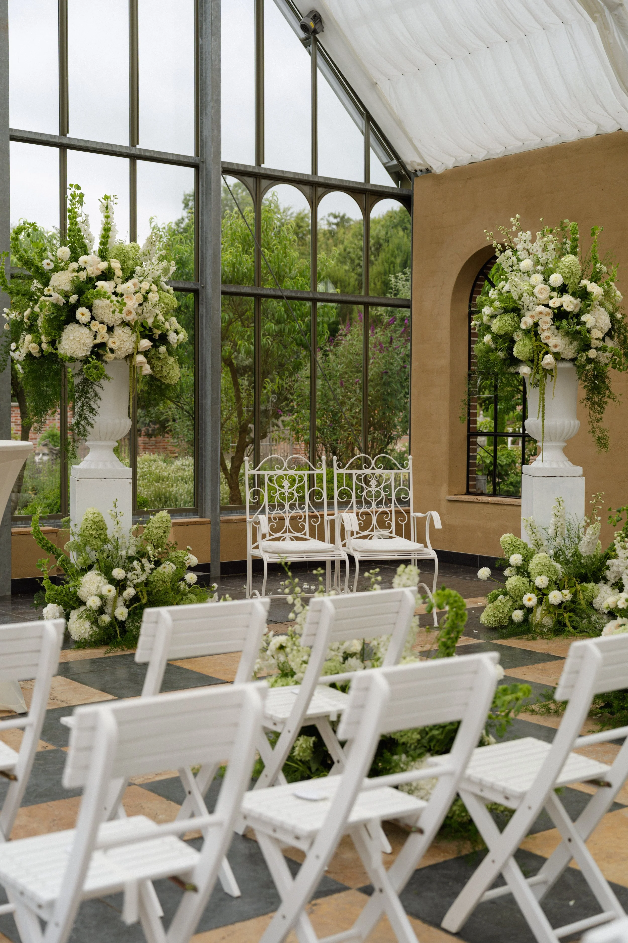 Innenraum einer Hochzeitsempfangshalle mit weißen Stühlen und großen Blumendekorationen in Vasen, große Fenster, Gartensicht im Hintergrund.