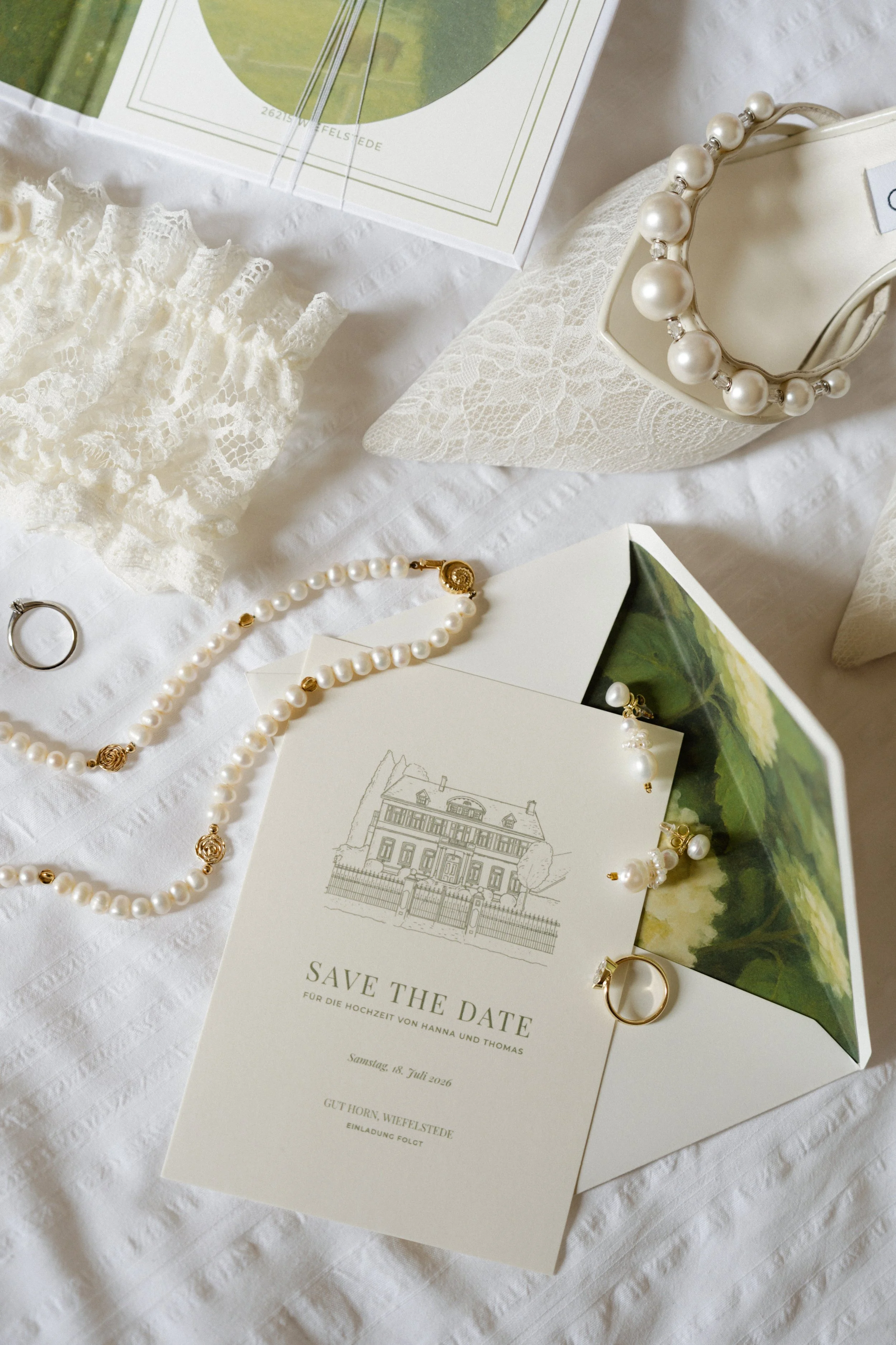 Hochzeitsankündigung mit Einladungskarte, weißer Perlenkette, Goldring, Spitzenaccessoire und Brosche auf weißem Stoff.
