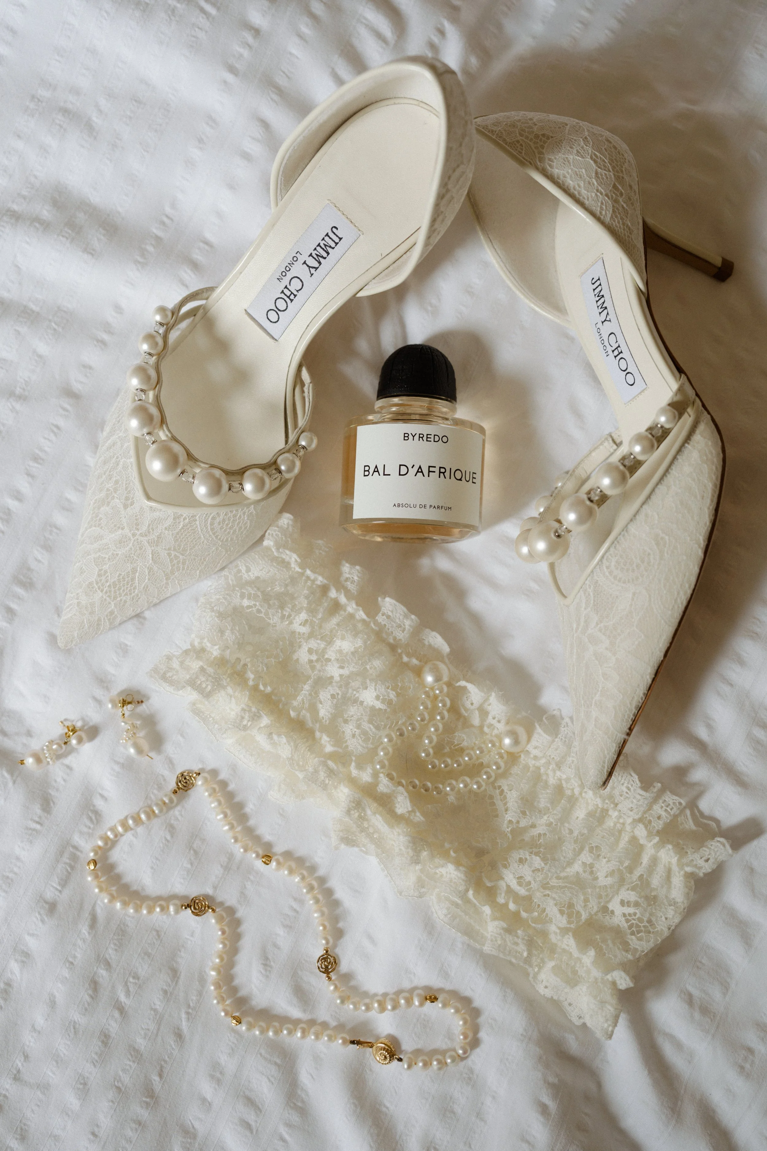 Man sieht ein Detailfoto der Accessoires der Braut. Brautschuhe von Jimmy Choo, ein Parfüm von Byredo, ein Strumpfband von Milla Nova, Ohre aus Perlen von ENA Studio.