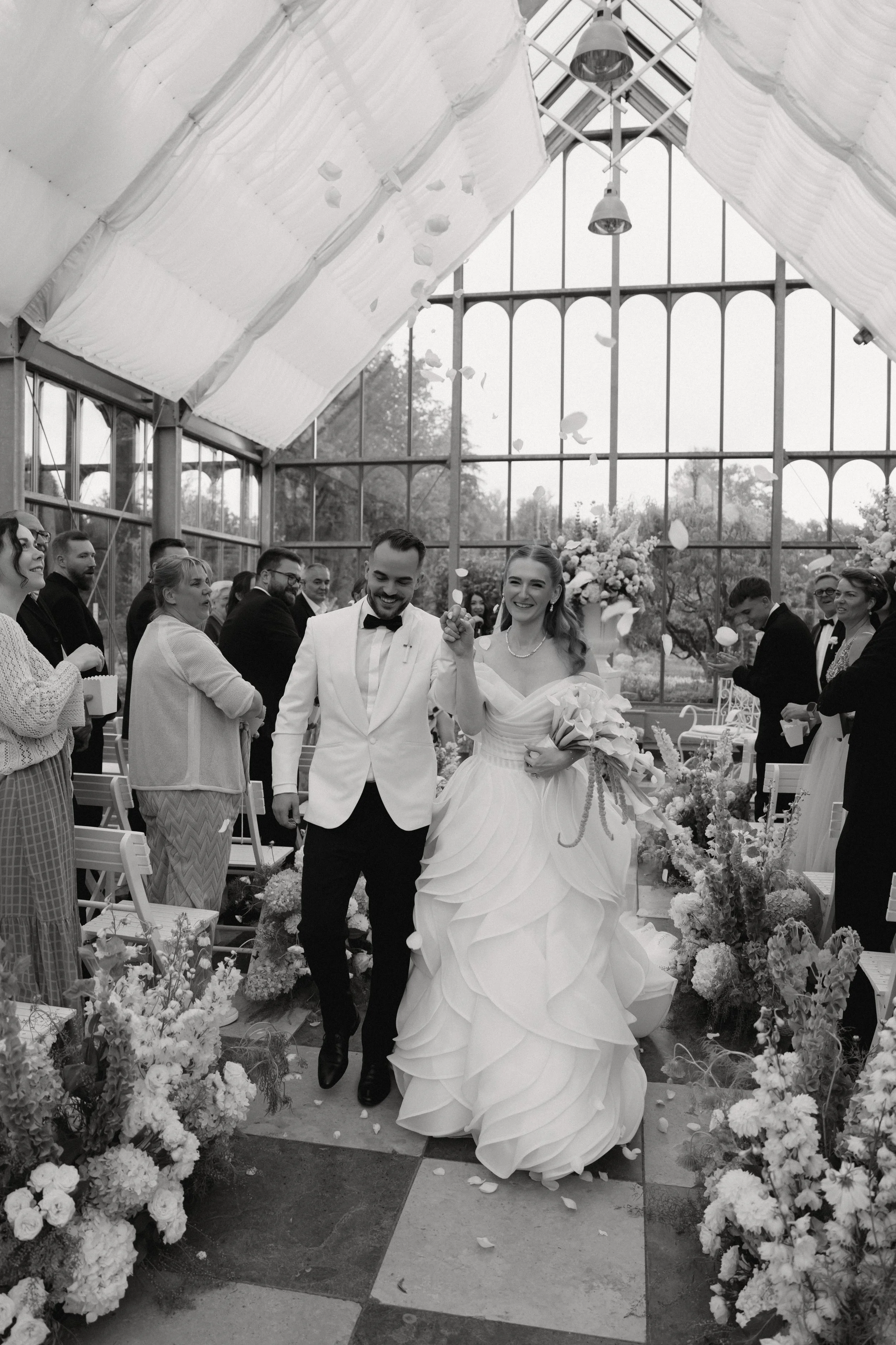 Ein Brautpaar bei einer Hochzeit, umgeben von Gästen, in einem lichtdurchfluteten Gewächshaus mit Blumen und Dekorationen.