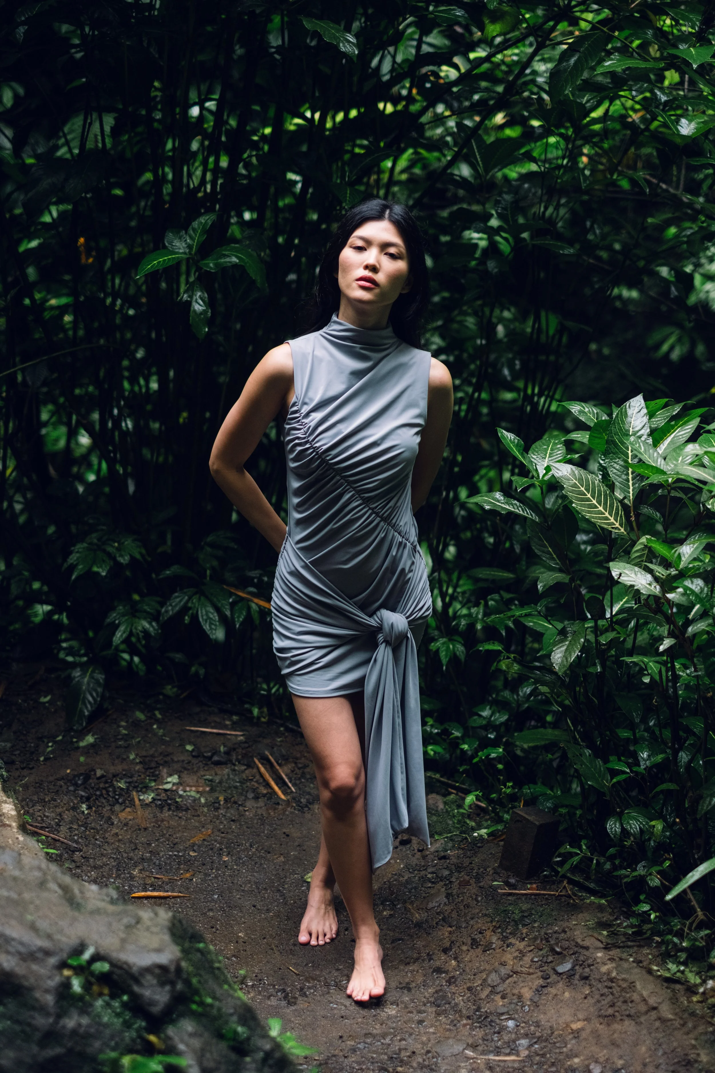 A woman in a gray dress standing barefoot on dark soil amidst lush green plants in a forest.