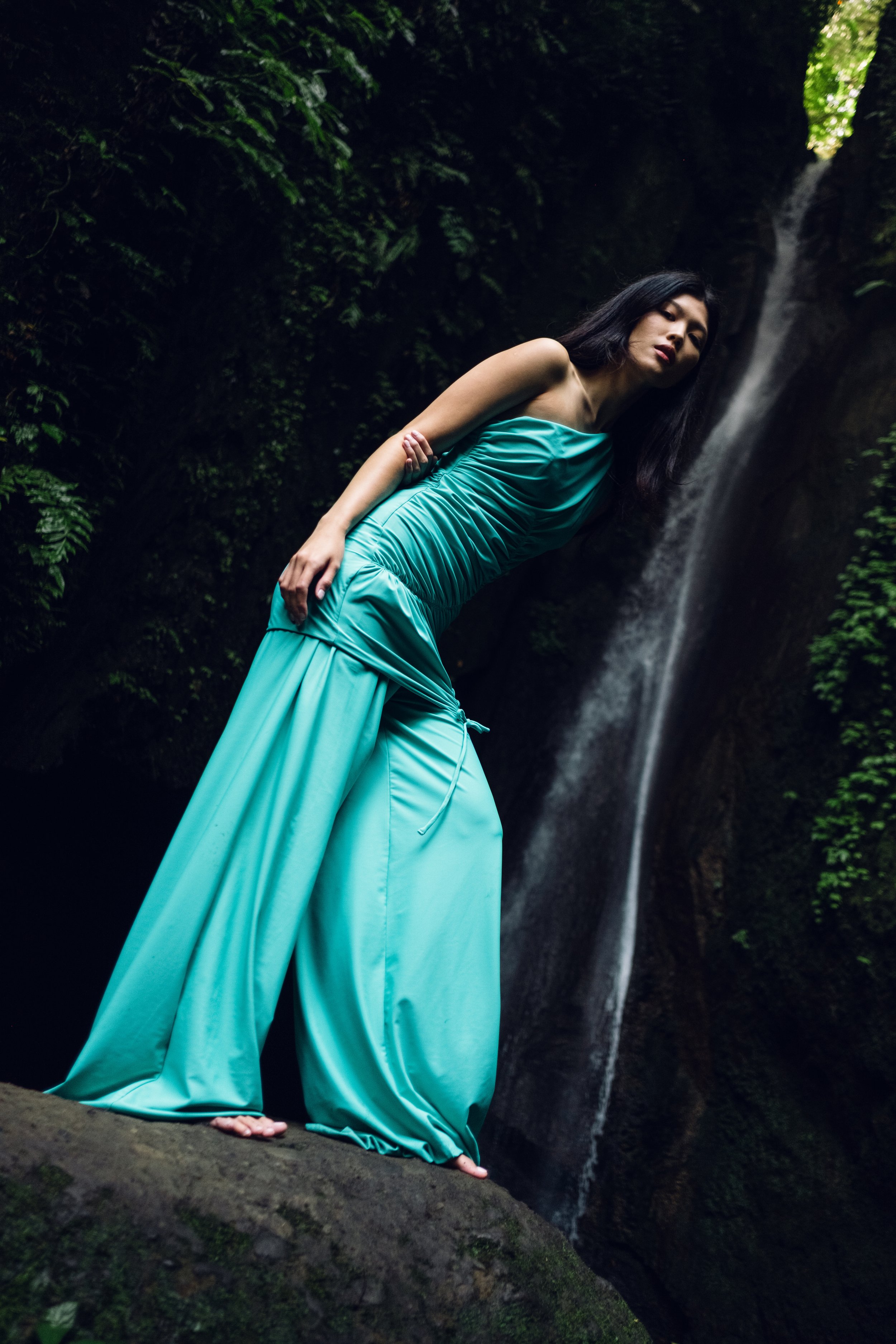 A woman in a flowing turquoise dress standing on rocks near a waterfall in a lush, green forest.