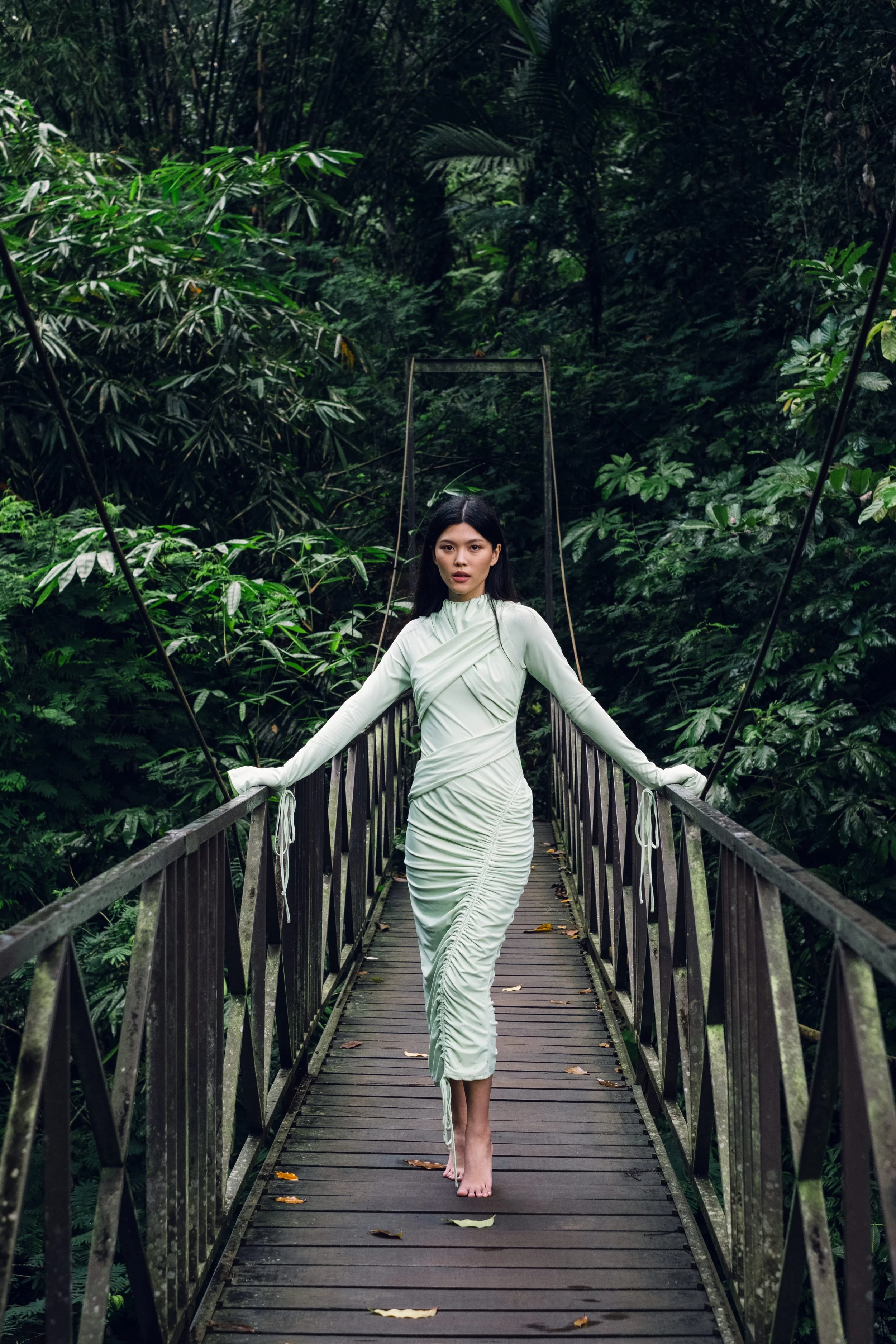A woman in a light-colored dress walking barefoot on a narrow wooden suspension bridge surrounded by lush green tropical foliage.