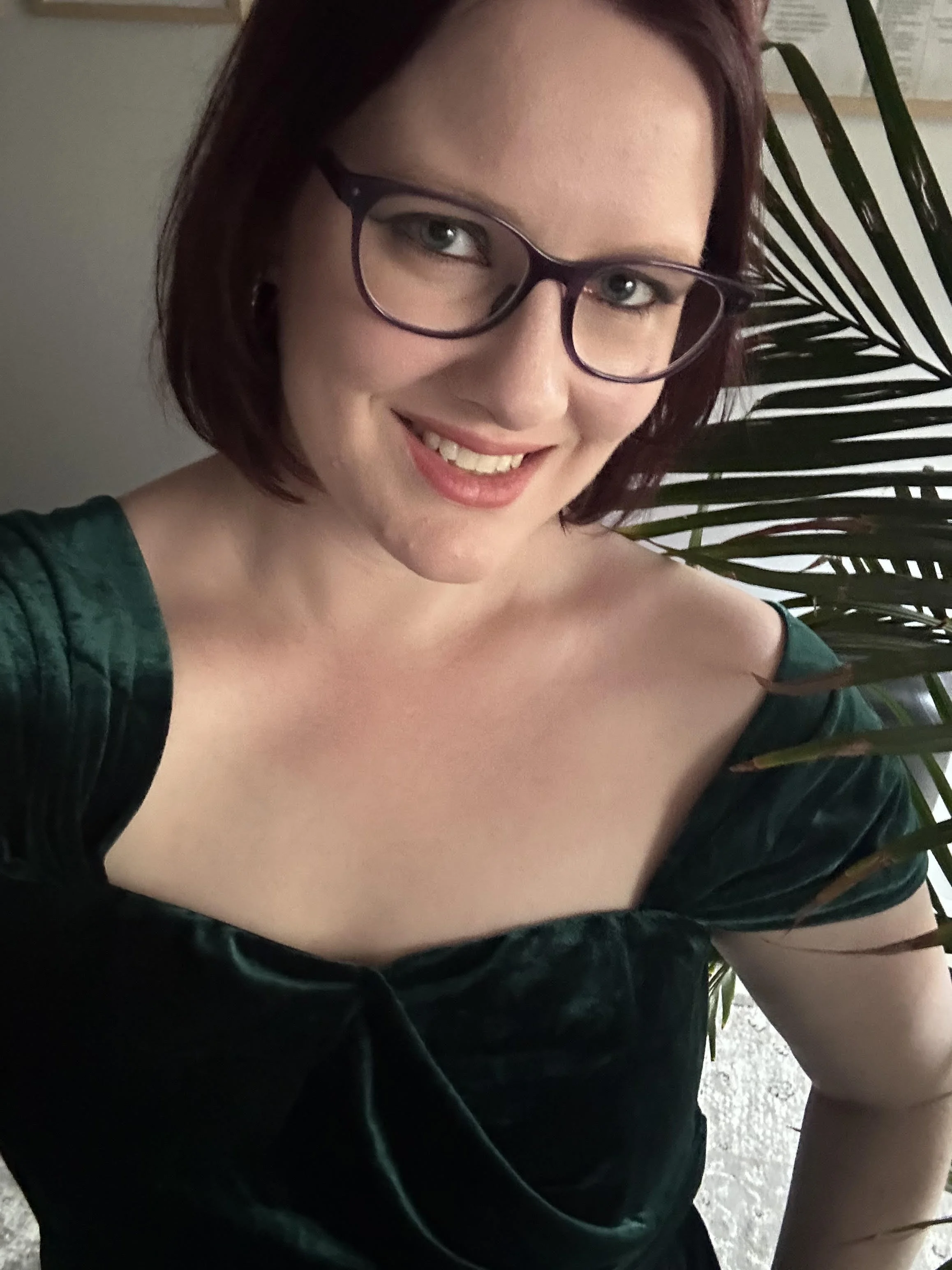 A woman with shoulder-length red hair and black glasses, smiling and wearing a dark green off-the-shoulder top, standing in front of a plant with long green leaves.