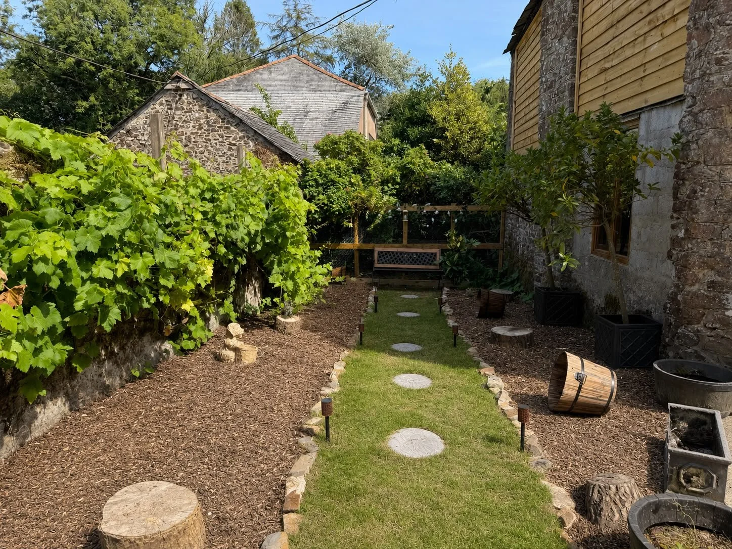A backyard garden with a pathway of stone slabs, surrounded by greenery, trees, and plants. There is a wooden bench at the end of the path, and various gardening items like pots and logs are visible on the sides for scent dog work. Treats area.