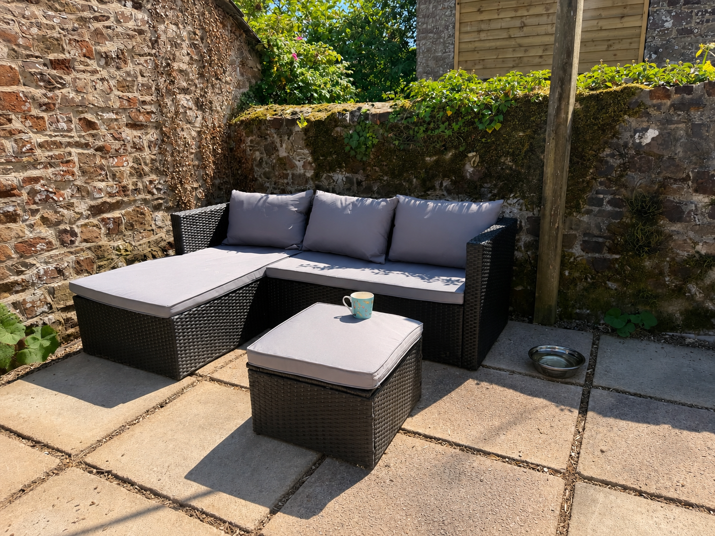 Outdoor patio with a black wicker sectional sofa with gray cushions, a matching ottoman with a coffee mug on top, a small metal water bowl, surrounded by a brick wall with some greenery and sunlight.
