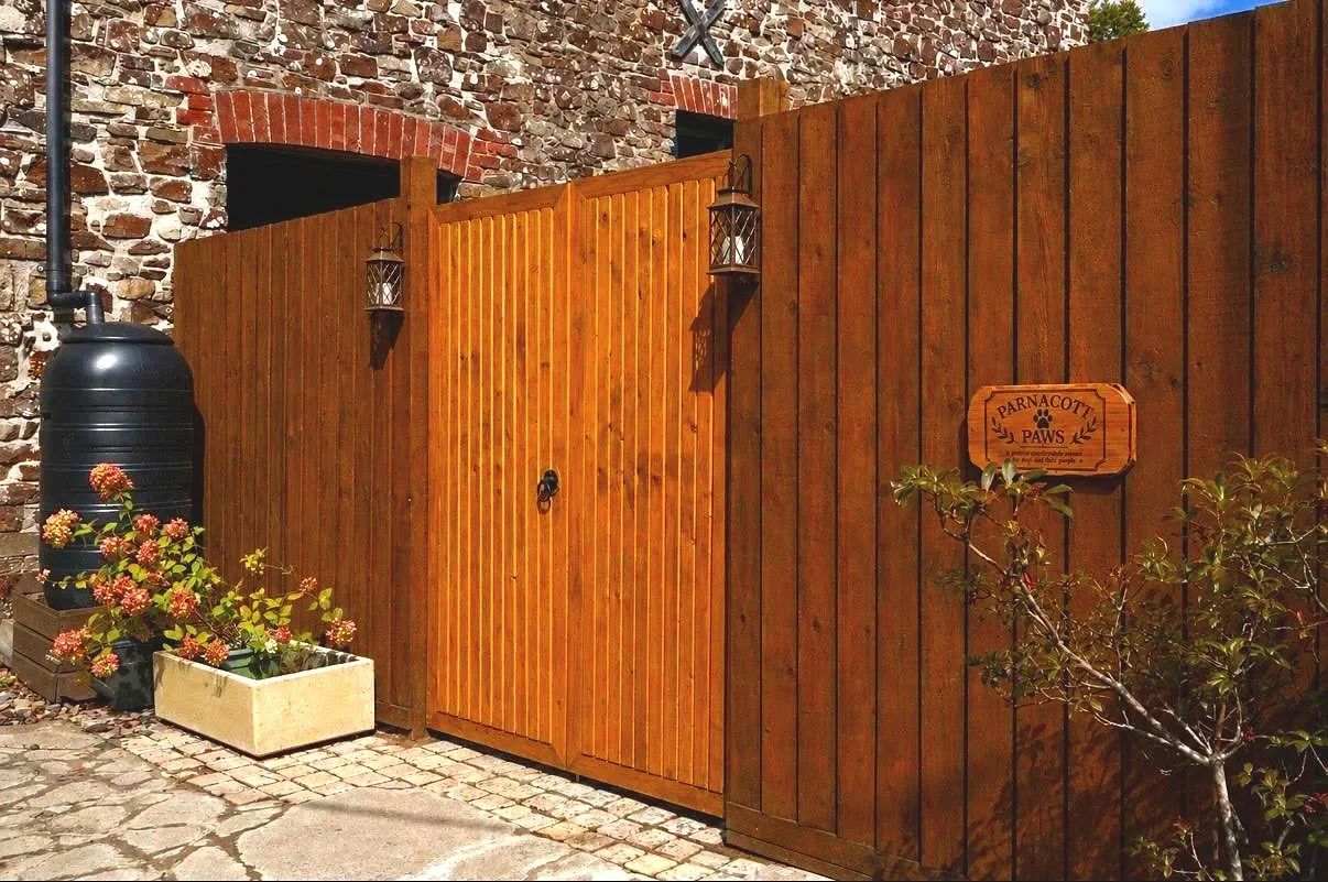 A wooden fence with a sign to the right that reads 'Parnacott Paws', an outdoor water container on the left, pink and yellow flowers in a white planter, and a brick wall in the background.