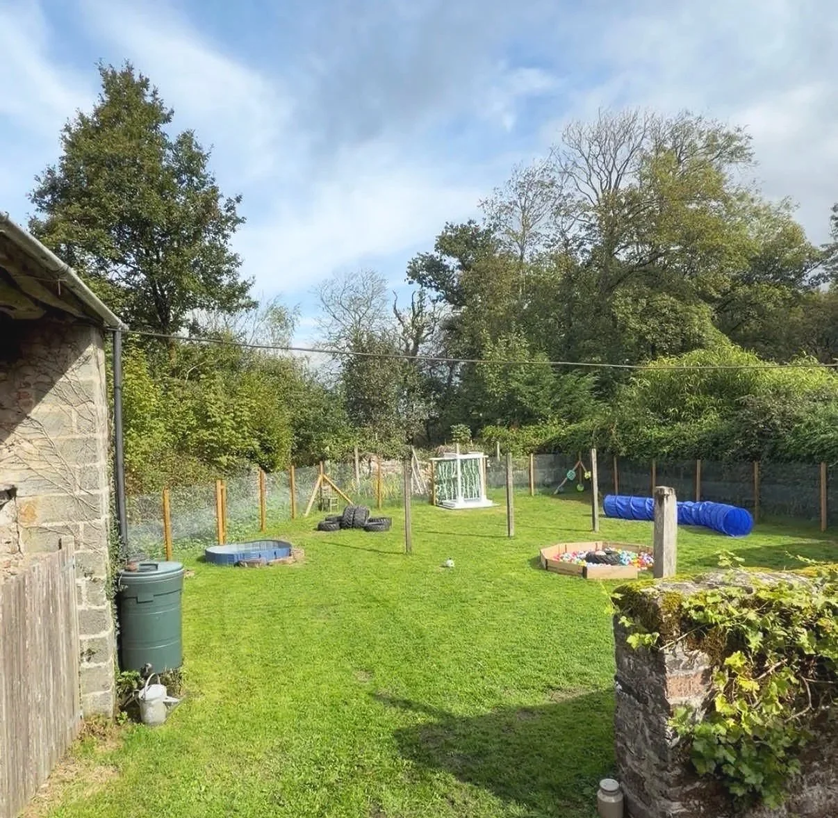 Parnacott paws overview with green grass, trees, a fence, and various outdoor play and garden equipment including a small pool, a tunnel, a sandbox, and a glass shed.