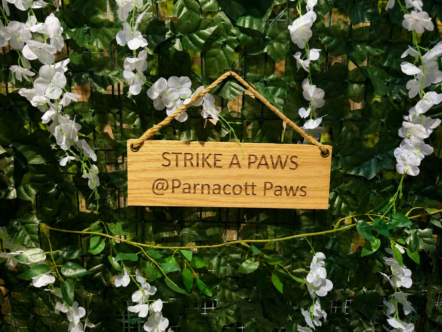 Wooden sign hanging on greenery with white flowers, reading 'Strike A Paws @Parnacott Paws'.