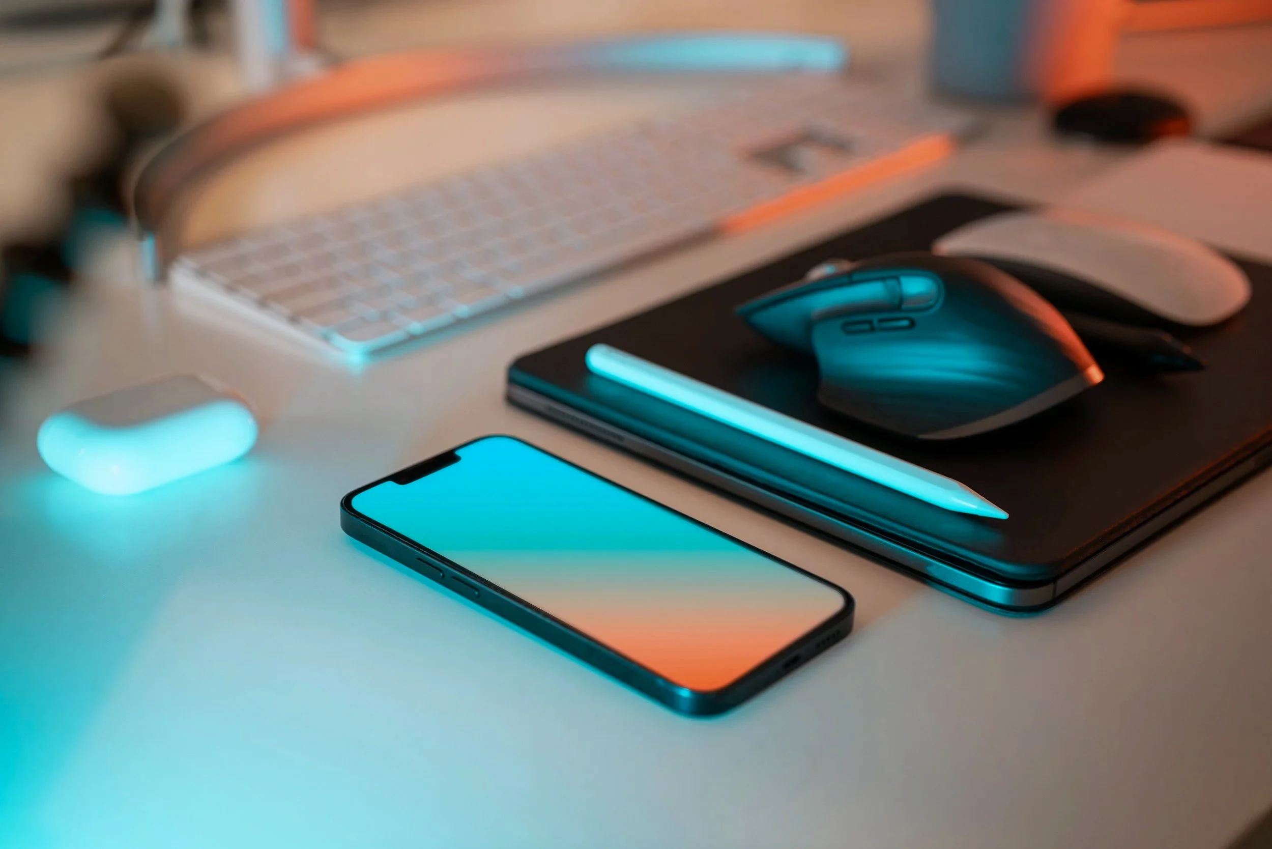 Ein Schreibtisch mit einem Smartphone, einem Laptop, einer Maus, Kopfhörern und einem Notizbuch, alles in warmem und kaltem Licht beleuchtet.