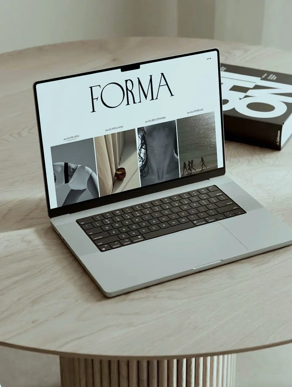 Laptop auf einem Holztisch zeigt eine Webseite mit dem Titel 'FORMA' und Bildern von Kunst und Natur, daneben ein Buch.