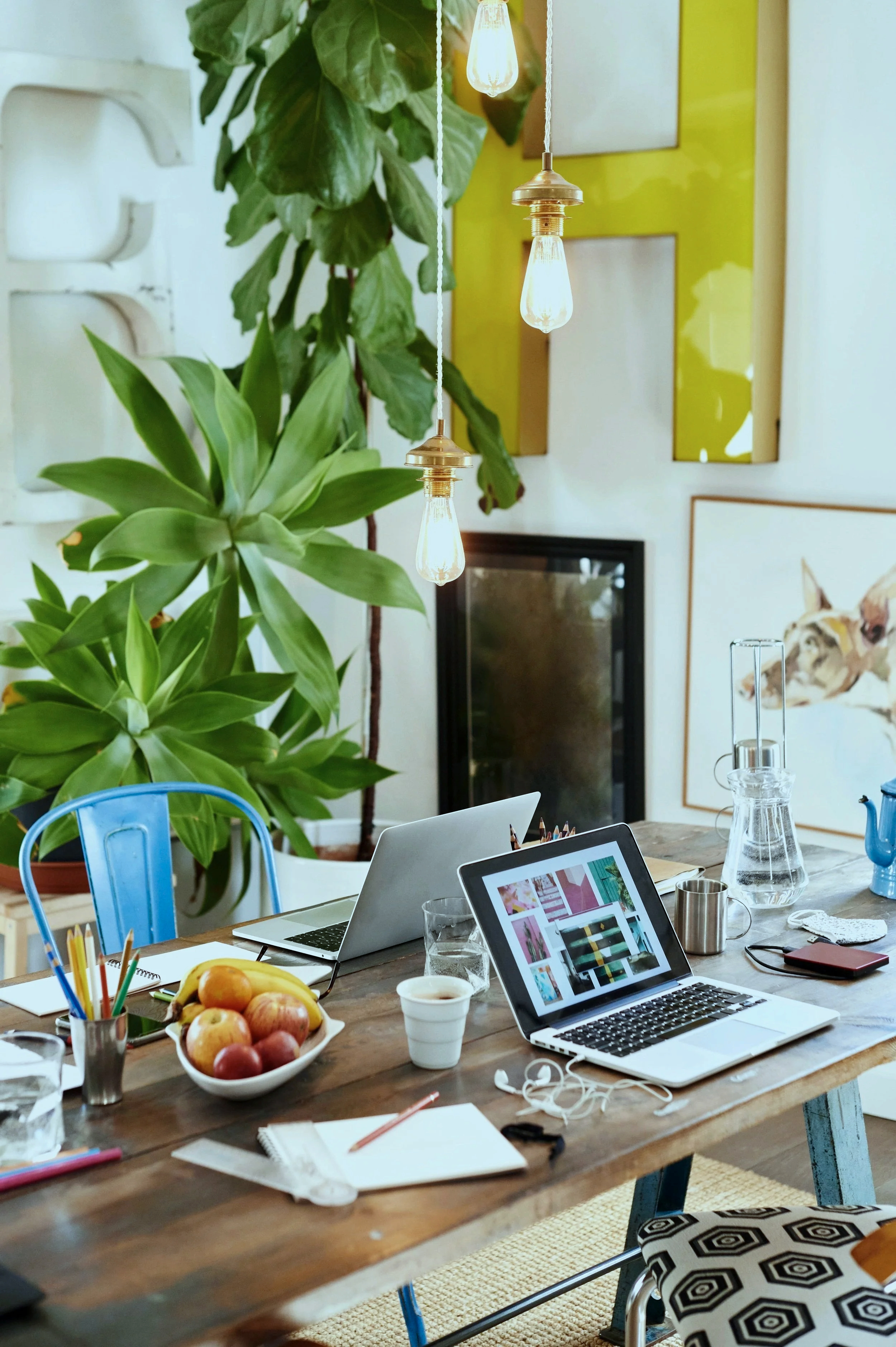 Ein großer Holztisch mit mehreren Laptops, einer Schale mit Äpfeln, Gläsern, Stiften, Notizbüchern, einem Notizblock und weiteren Gegenständen. Im Hintergrund sind große grüne Zimmerpflanzen, Wanddekoration und eine Lampe mit sichtbarer Glühbirne. Ein Sitz mit dekorativem Sitzkissen.