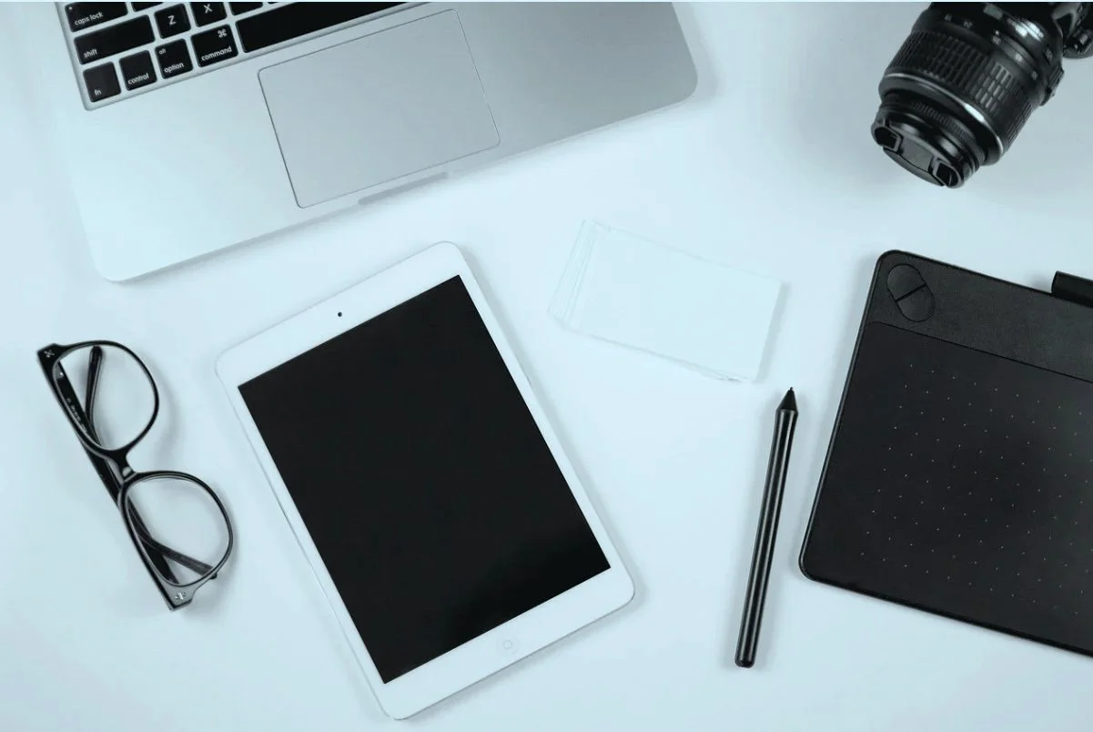 Arbeitsplatz mit Laptop, Tablet, Brille, Kamera, Stift, Notizblock, Speicherkarte, und Zeichenpad auf weißem Tisch.